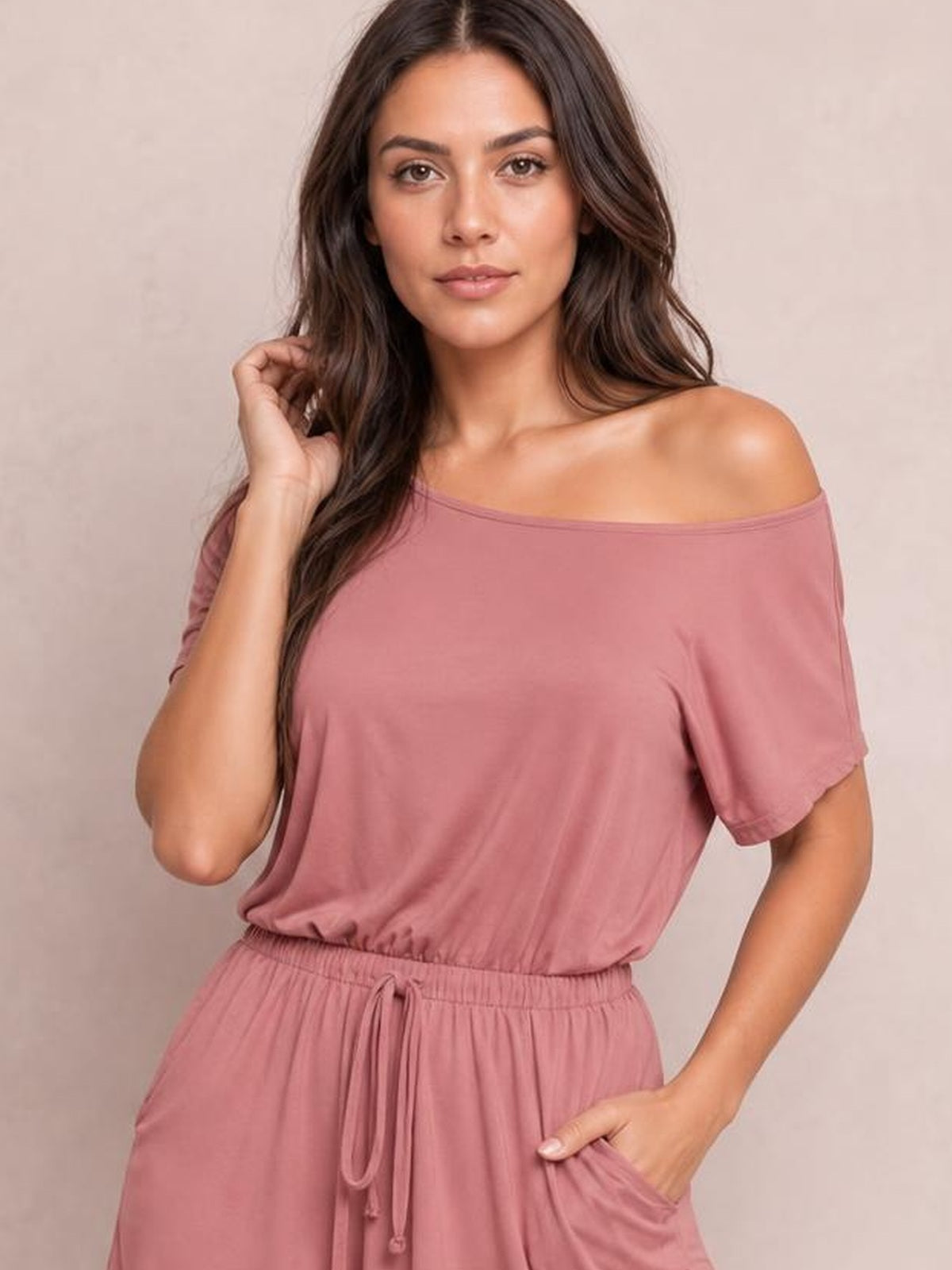 Model wearing a dusty rose short-sleeve romper with a wide boat neckline, elastic drawstring waist, relaxed silhouette, and practical side pockets.