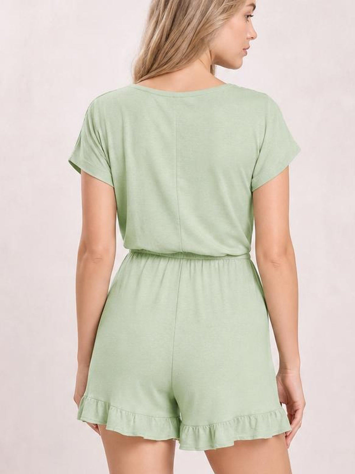 A model stands turned away in a mint green ruffled boat neck short sleeve romper with an elastic waist, relaxed silhouette, and flounced shorts hem.