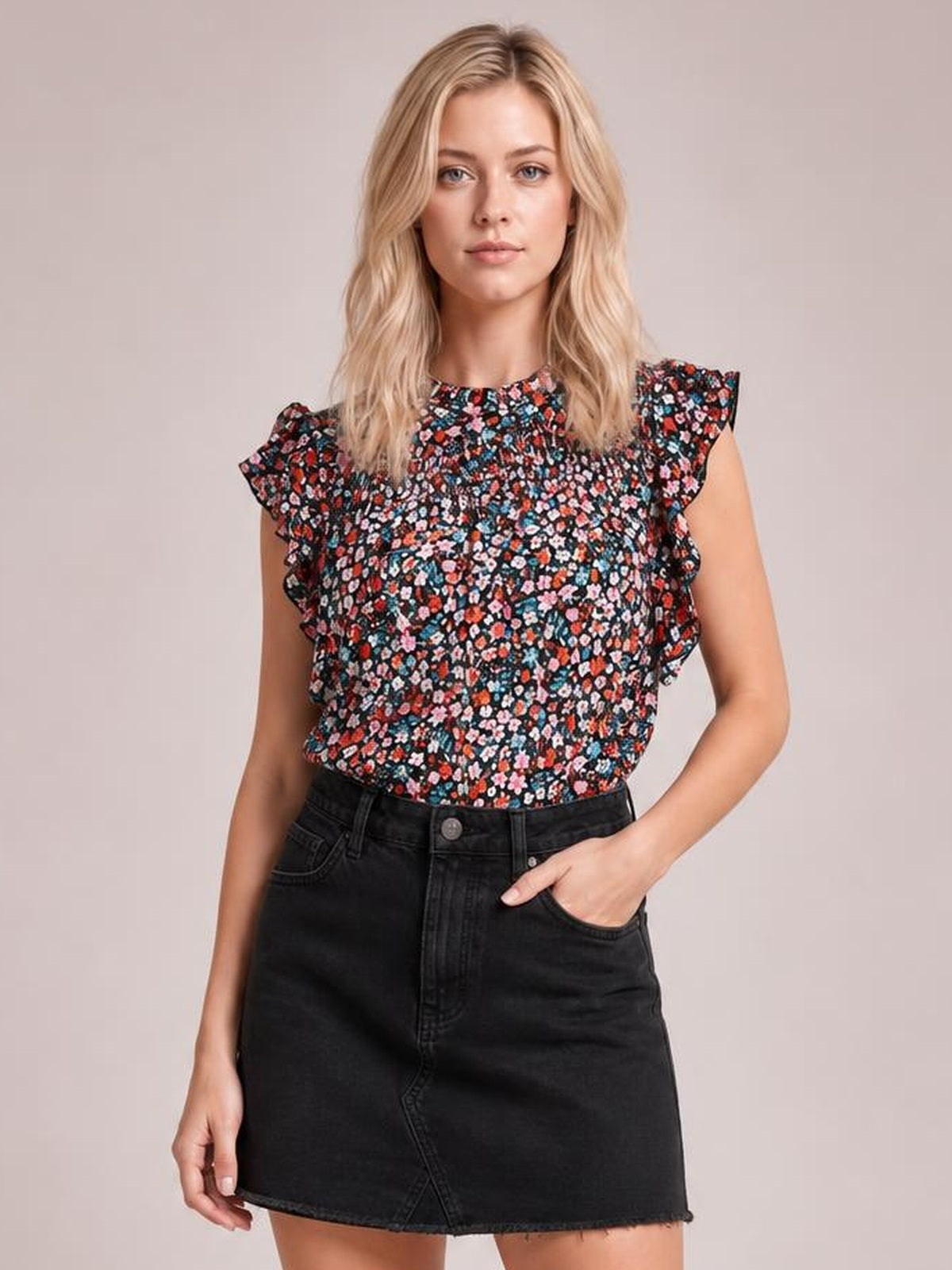 A model stands with one hand in her pocket, wearing a black ditsy floral blouse with a mock neck, ruffled cap sleeves, and a softly smocked silhouette.