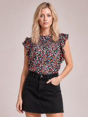 A model stands with one hand in her pocket, wearing a black ditsy floral blouse with a mock neck, ruffled cap sleeves, and a softly smocked silhouette.