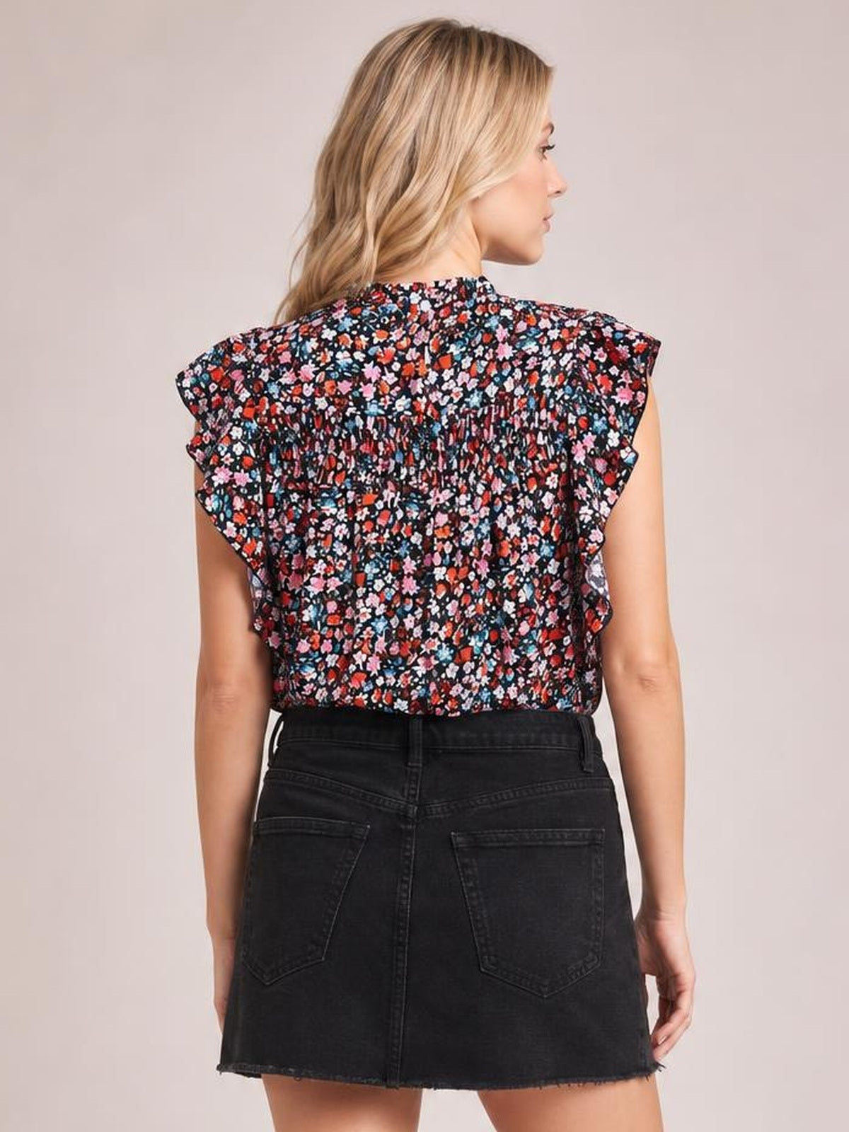 A model stands facing away in a black ditsy floral blouse with a mock neck, ruffled cap sleeves, and a smocked back yoke.