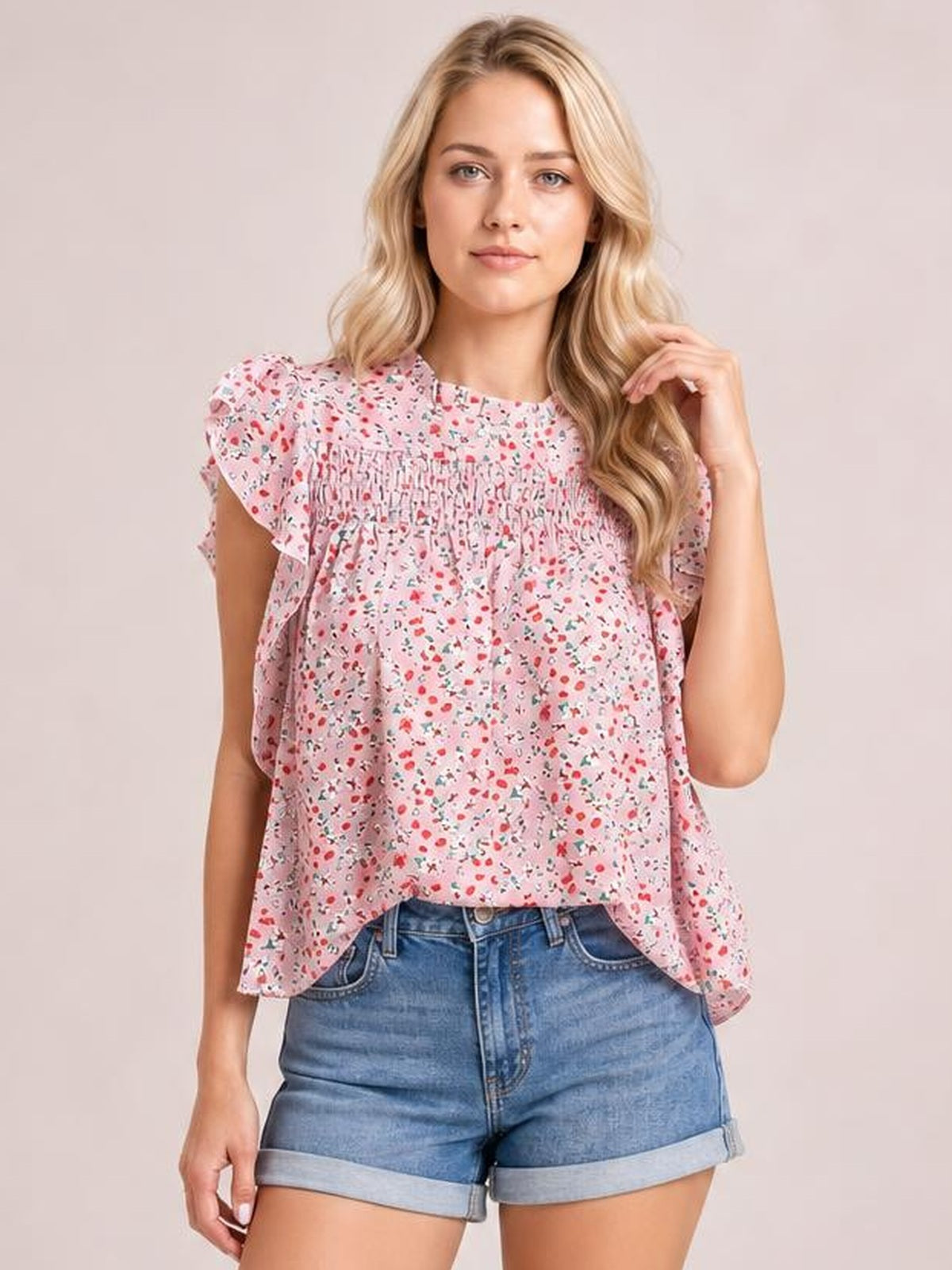 Model poses in a pink ditsy floral blouse with a mock neck, ruffled cap sleeves, smocked yoke, and relaxed silhouette.