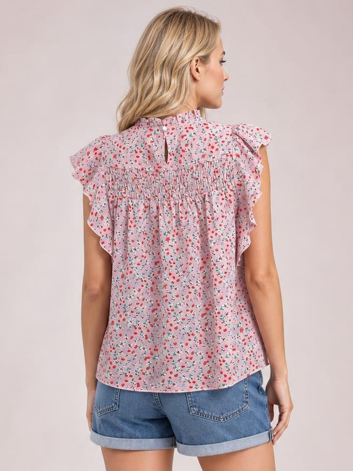 A model faces away in a pink ditsy floral blouse with a ruffled mock neck, cap sleeves, smocked back panel, and relaxed straight silhouette.