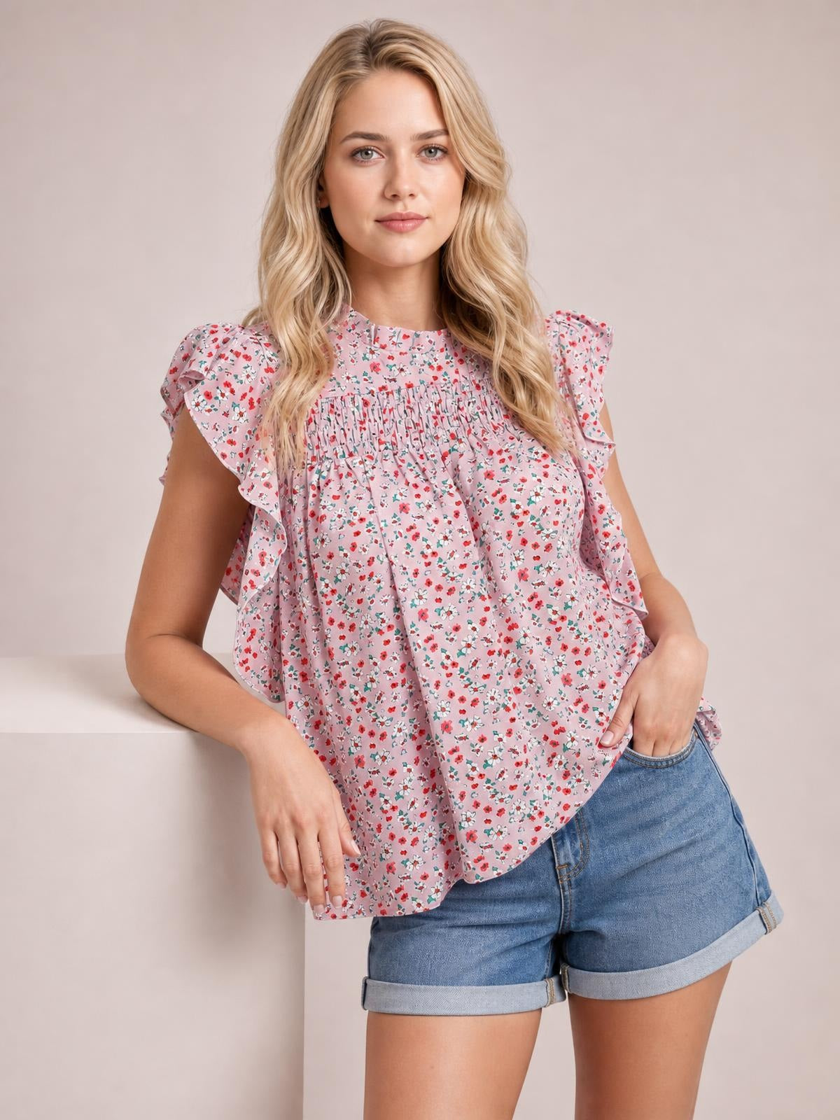 Model poses with one hand in her pocket wearing a pink ditsy floral blouse with a mock neck, ruffled cap sleeves, smocked yoke, and relaxed silhouette.