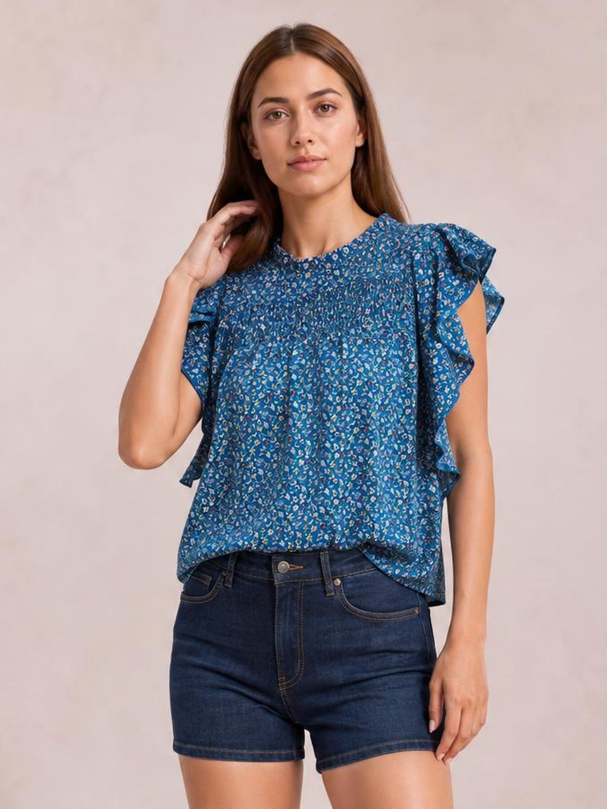 A woman poses with one hand in her hair wearing a blue ditsy floral blouse with a mock neck, smocked bodice, ruffled cap sleeves, and a relaxed silhouette.