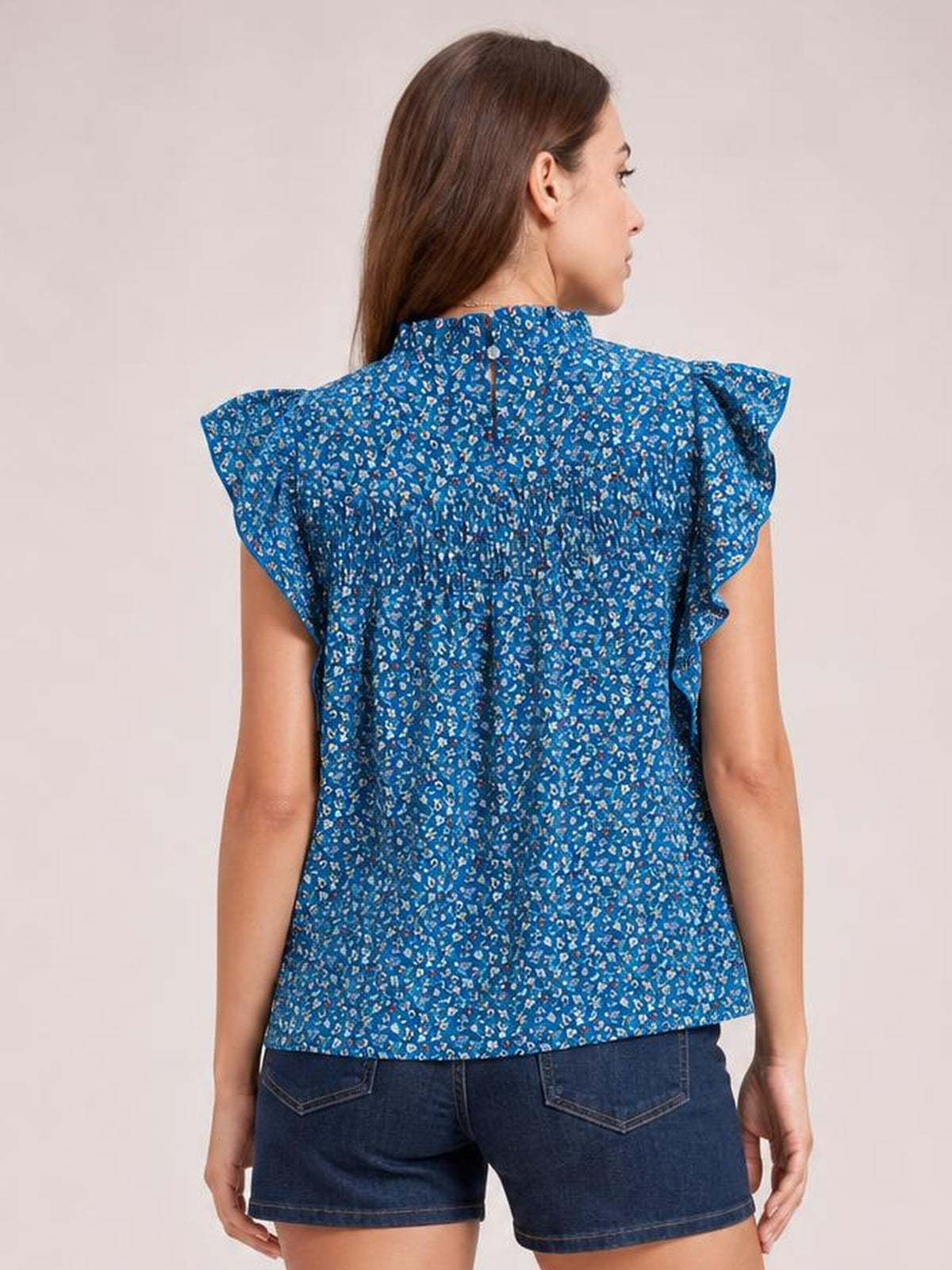 Model shown from the back in a blue ditsy floral blouse with a smocked bodice, ruffled cap sleeves, and a high mock neck, turning to the side.