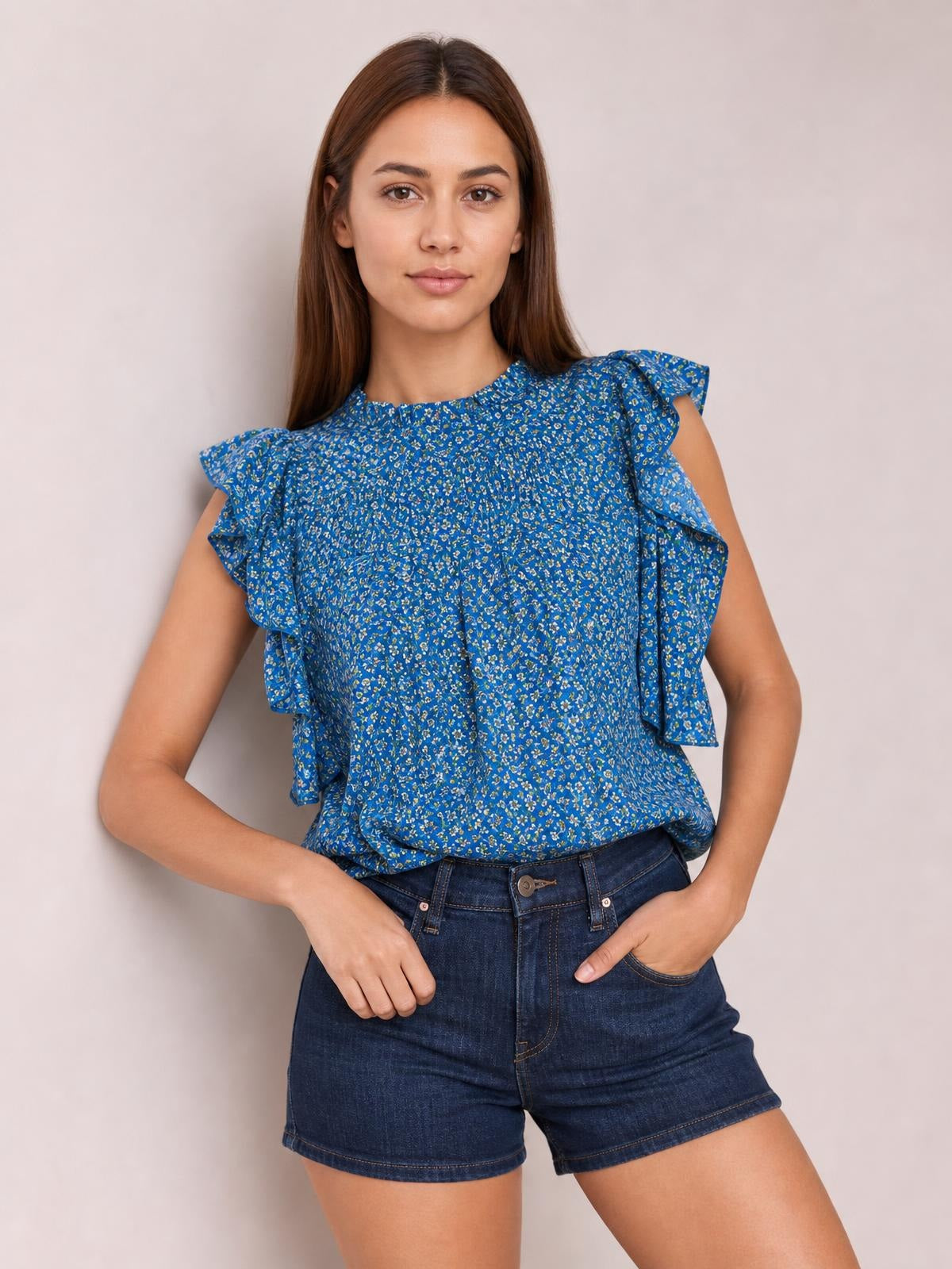 Model wearing a blue ditsy floral blouse with a smocked bodice, ruffled cap sleeves, and a softly gathered mock neck, paired with denim shorts.