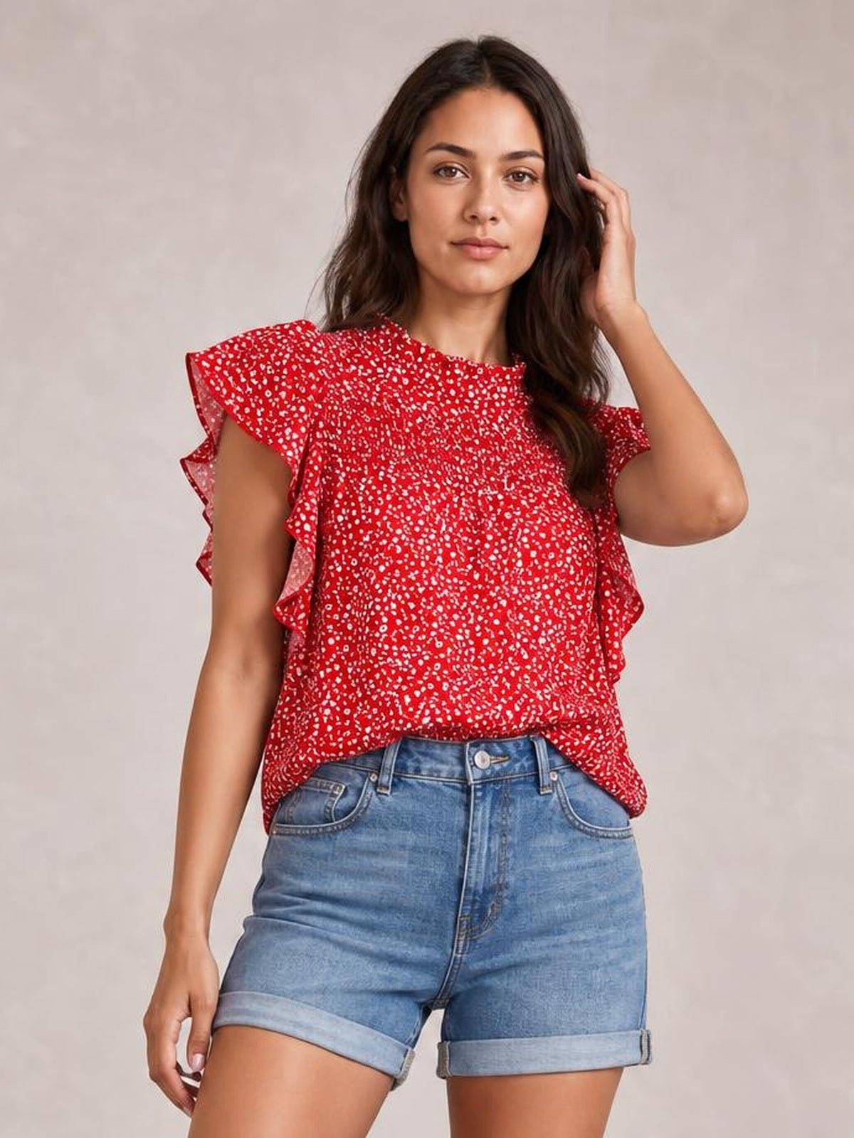 Model wearing a red ditsy floral blouse with a mock neck, ruffled cap sleeves, smocked texture, and a relaxed silhouette, touching her hair.