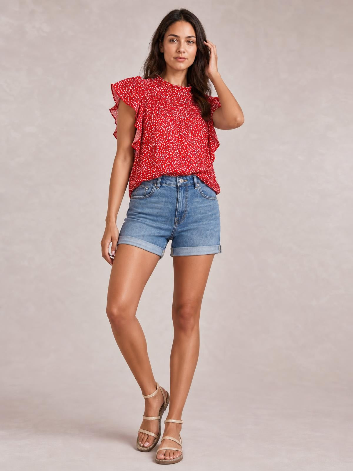 Model stands with one hand touching her hair, wearing a red ditsy floral blouse with a mock neck, smocked bodice, cap sleeves, and ruffled frill edges.