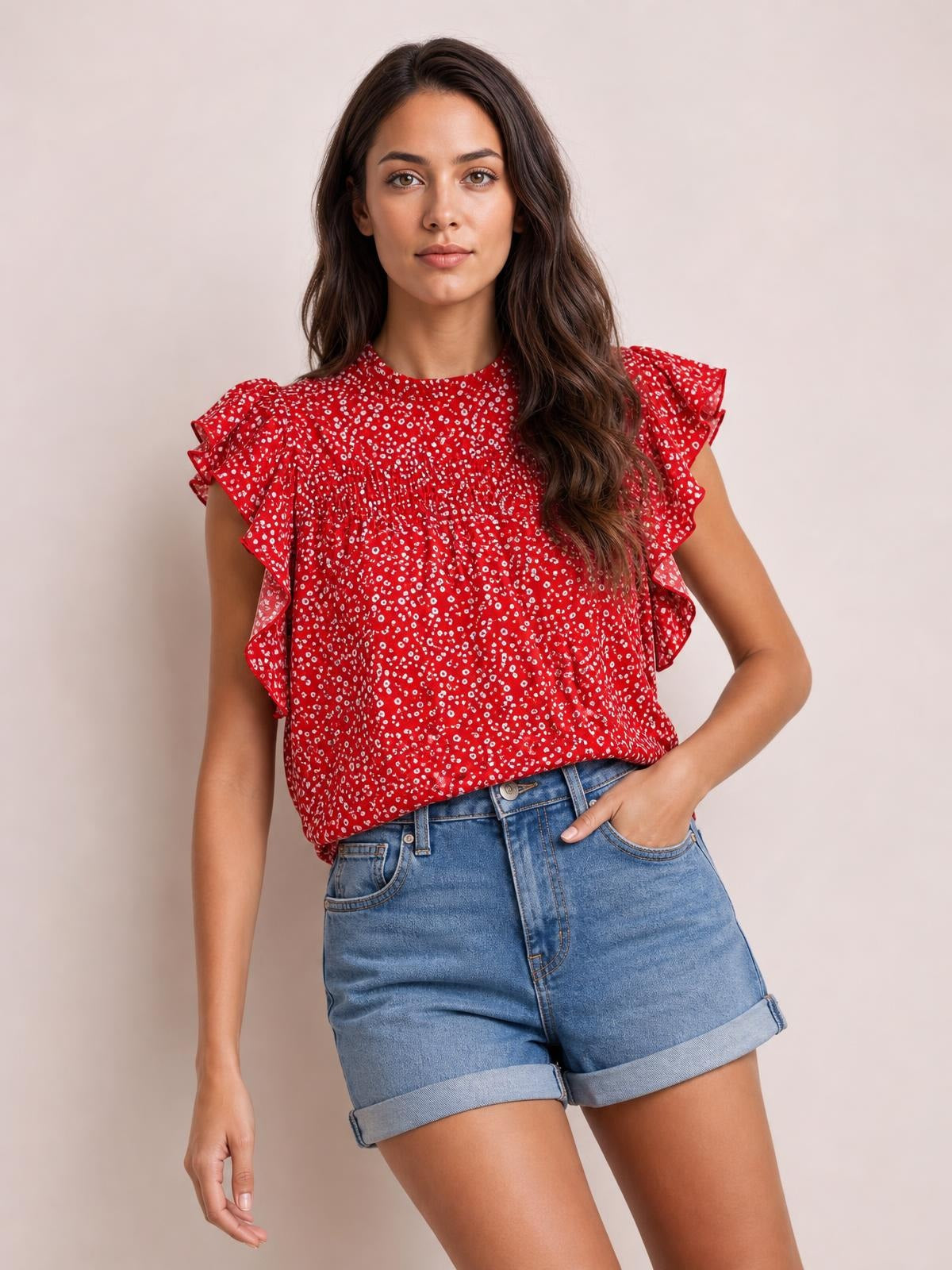 A model poses with one hand in her pocket wearing a red ditsy floral blouse with a mock neck, ruffled cap sleeves, smocked bodice, and relaxed silhouette.
