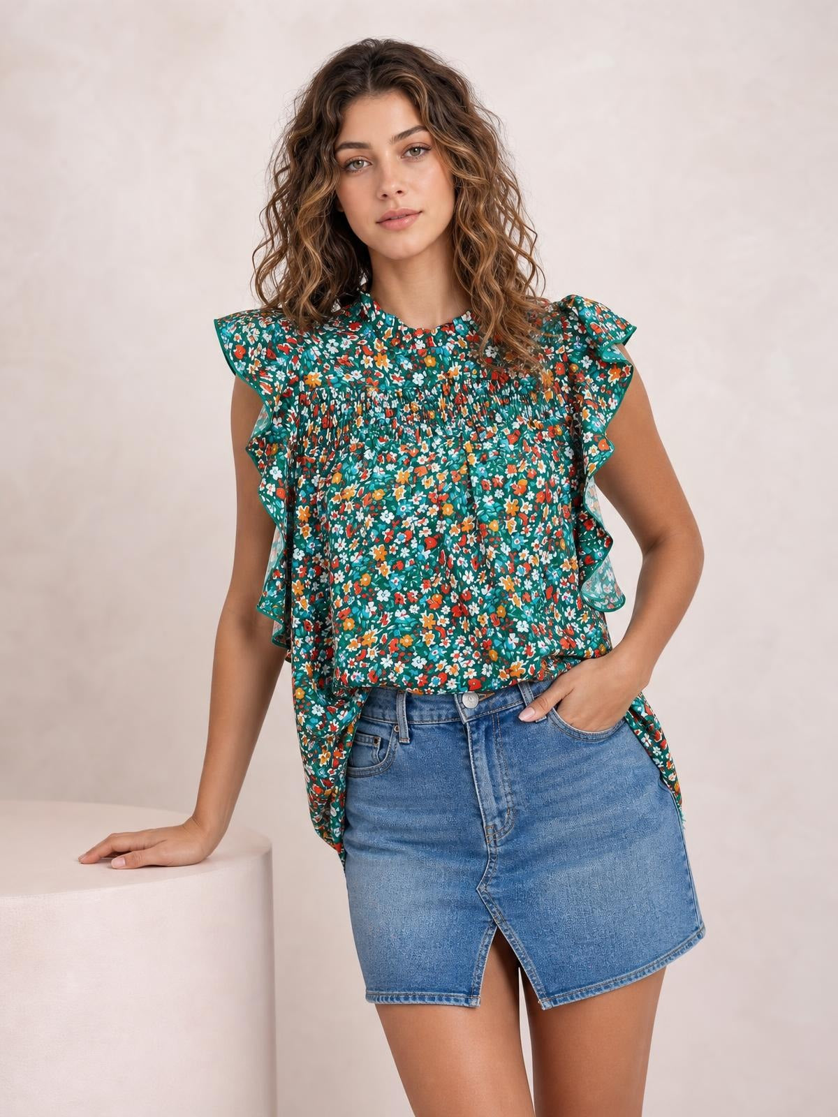 A model poses in a teal ditsy floral blouse with a ruffled mock neck, fluttering cap sleeves, smocked bodice, and relaxed flowing silhouette.