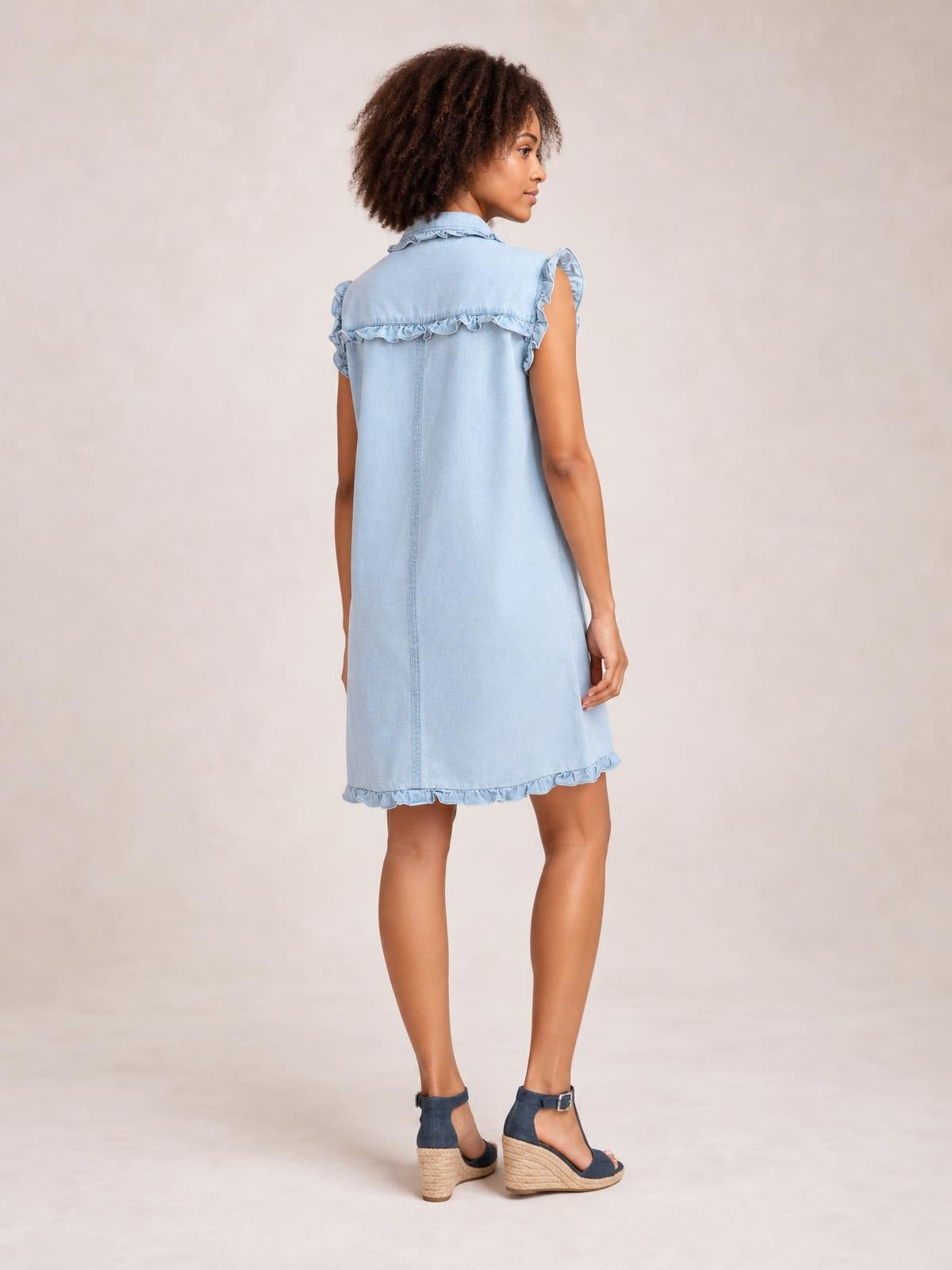 Model shown from behind in a light blue denim shift dress with a ruffled collar, sleeveless ruffled edges, center seam, and ruffled hem.