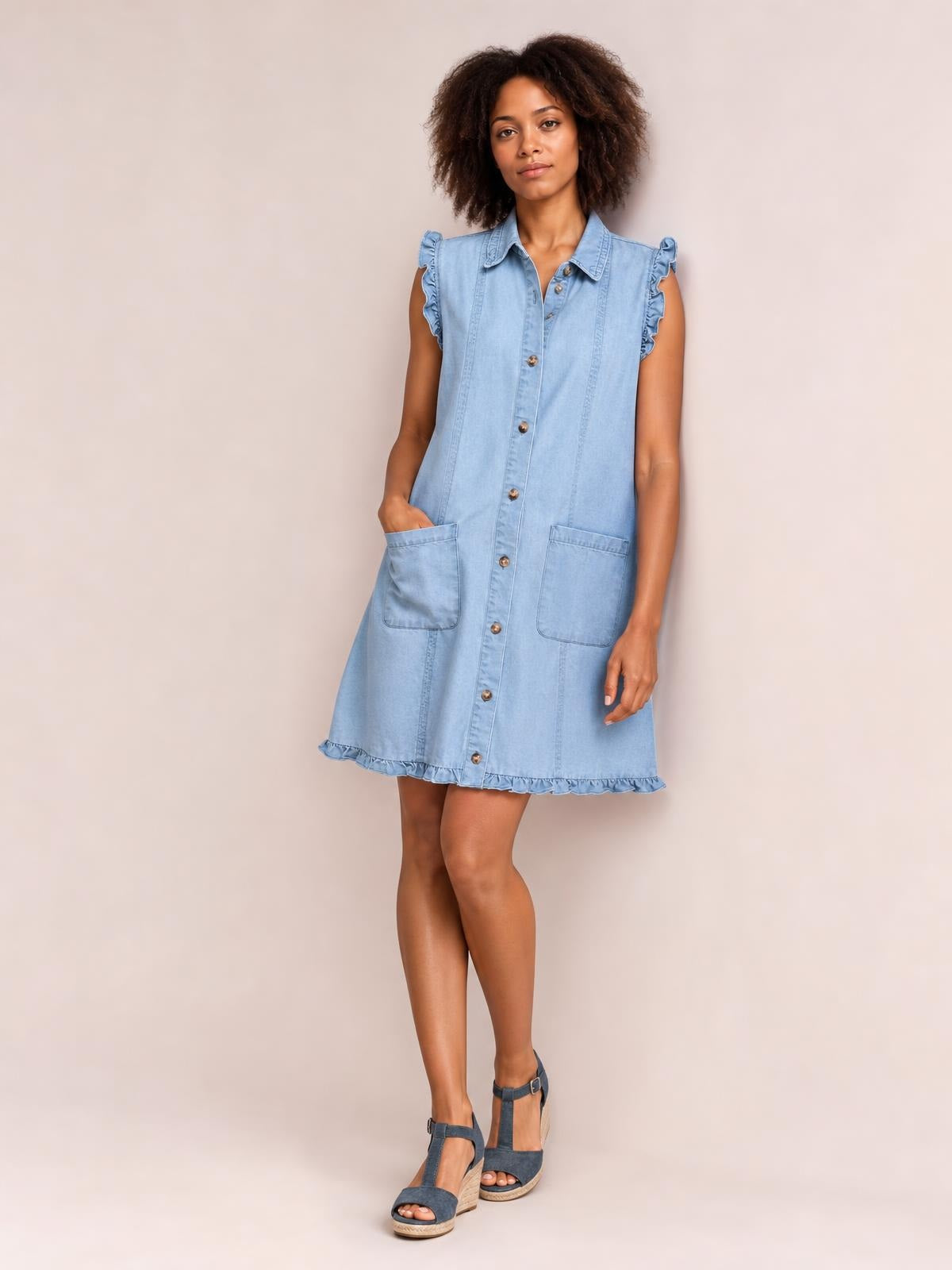 Model wears a light blue denim shift dress with a collar, button-front construction, ruffled sleeveless edges and hem, patch pockets, and a relaxed silhouette.