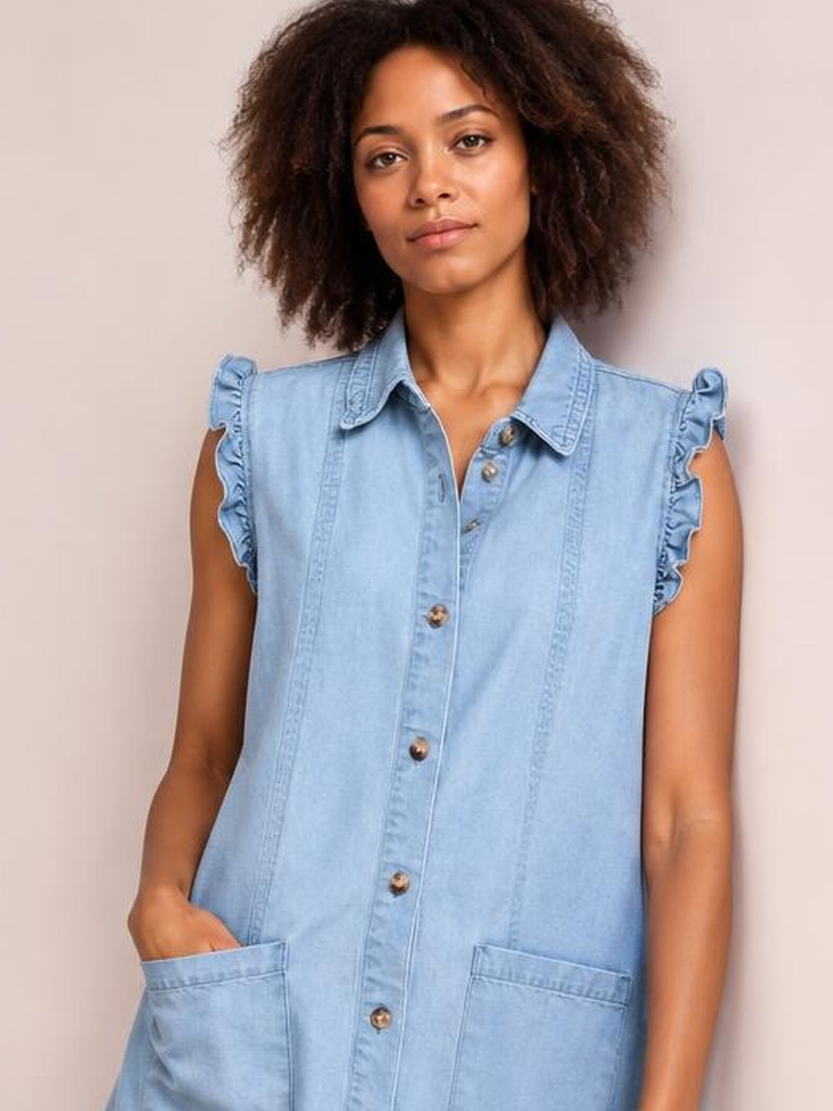 Model poses with one hand in a pocket, wearing a light blue denim shift dress with a collared button front, ruffled cap sleeves, and patch pockets.
