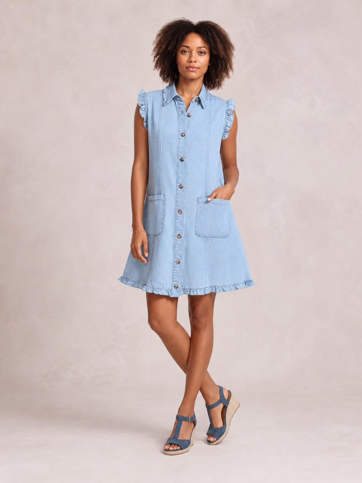 Model in a light blue denim shift dress with a button-front collared design, ruffled cap sleeves and hem, two patch pockets, and a flared silhouette.