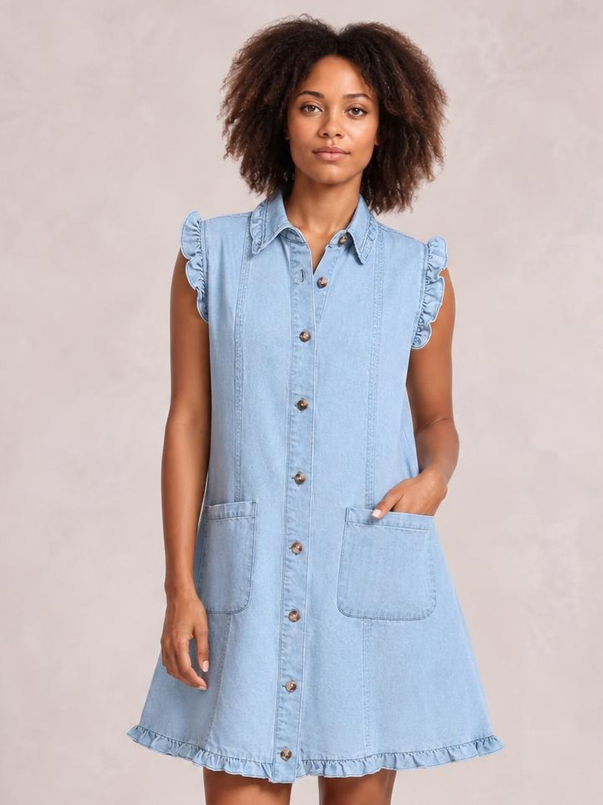 Model wears a light blue denim shift dress with a collared button front, ruffled cap sleeves and hem, large patch pockets, and a relaxed A-line silhouette.