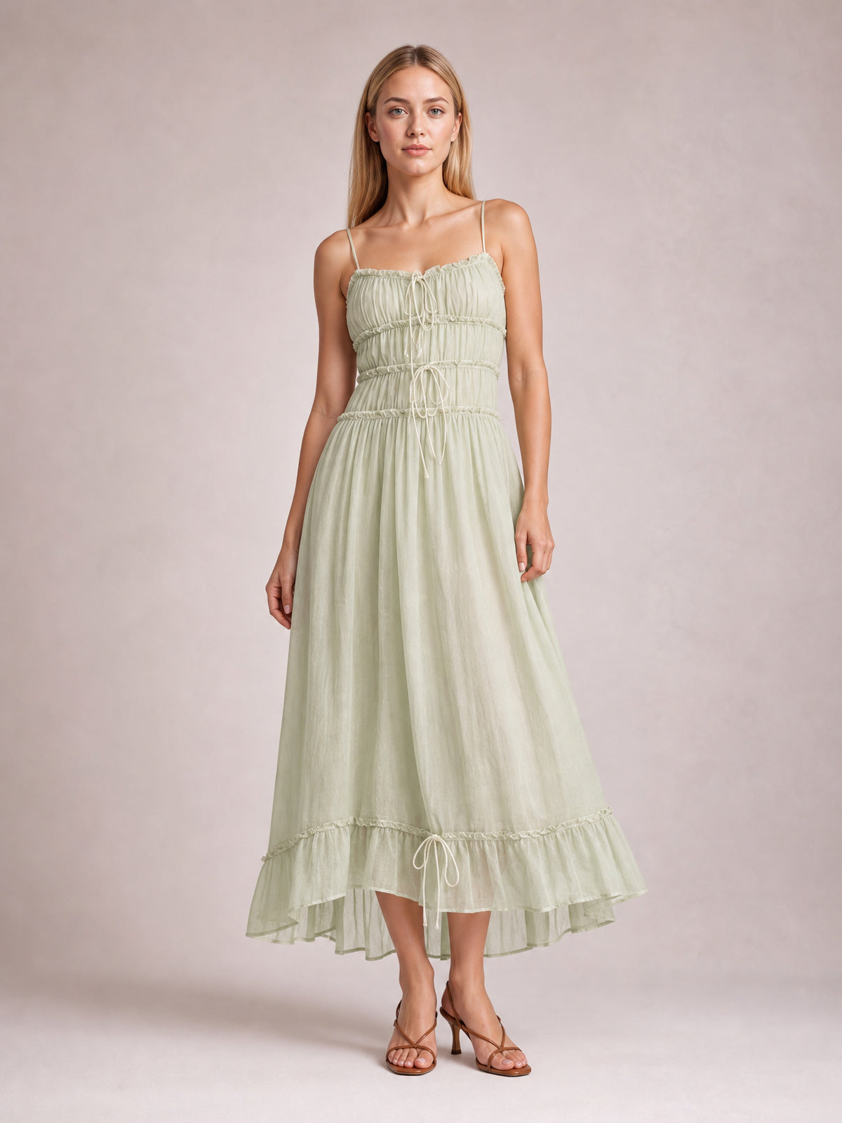Model wearing a light green ruffled hem frill midi cami dress with spaghetti straps, a gathered bodice, front tie details, and a flowing tiered silhouette.