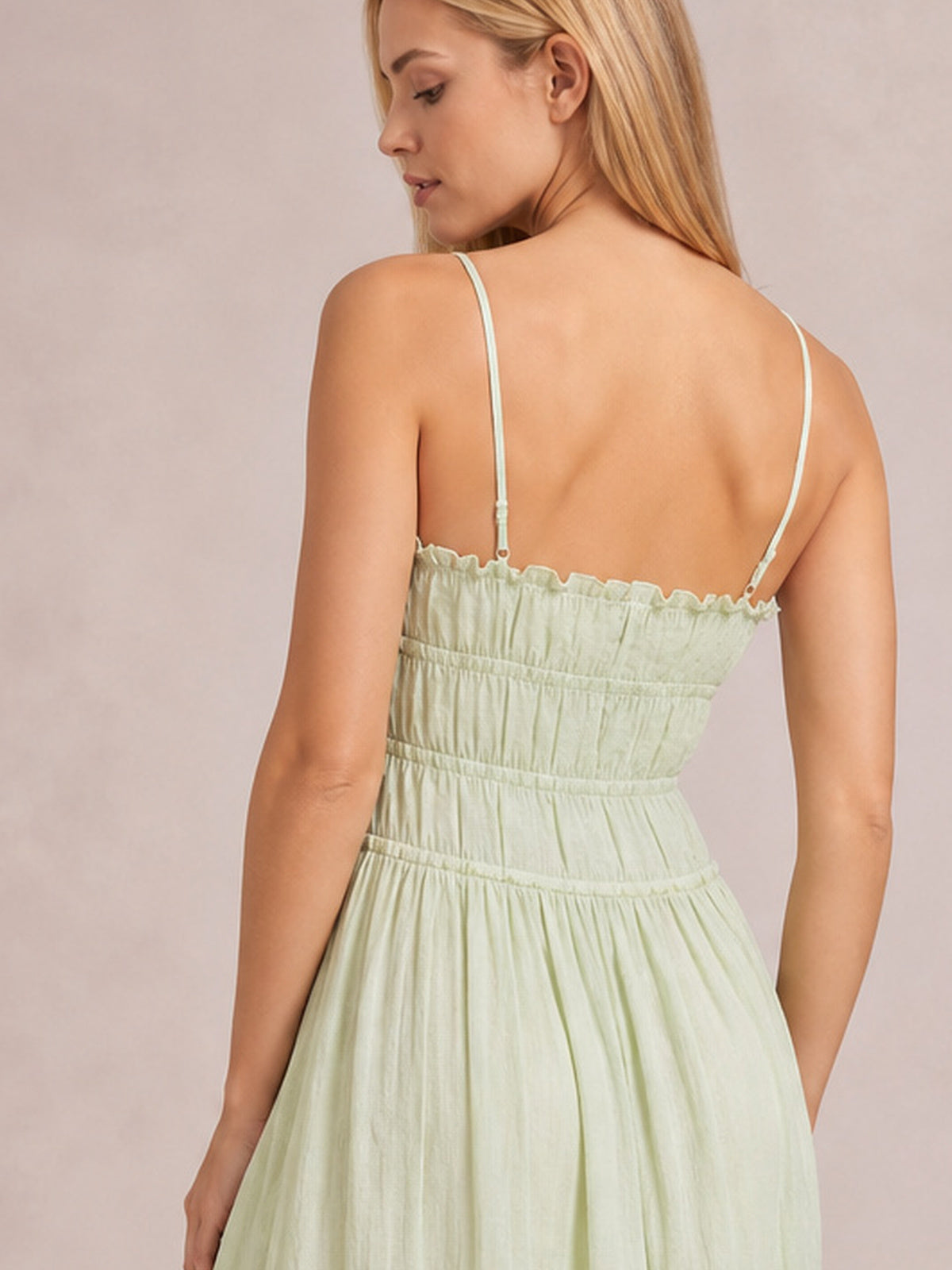 A model turns away in a light green cami midi dress with thin spaghetti straps, a gathered back bodice, defined waist, and fluid, full skirt silhouette.