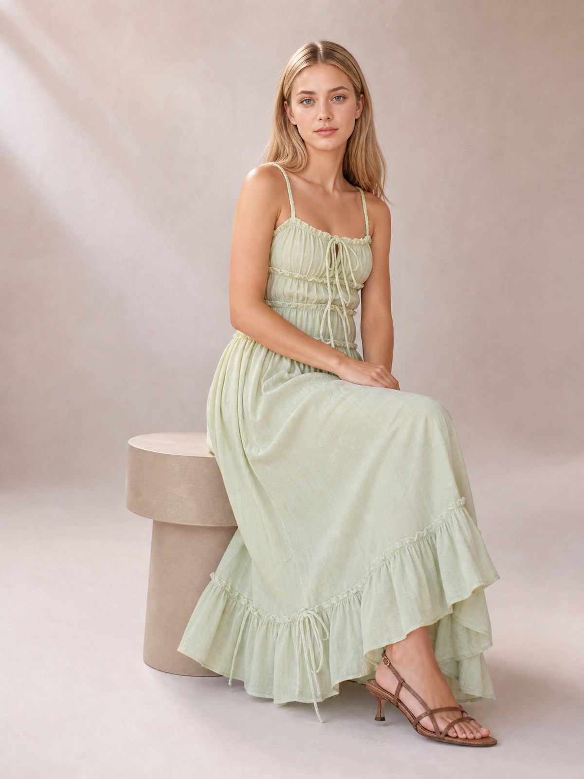 A seated model wears a light green midi cami dress with spaghetti straps, a softly gathered tied bodice, flowing silhouette, and layered ruffled hem.