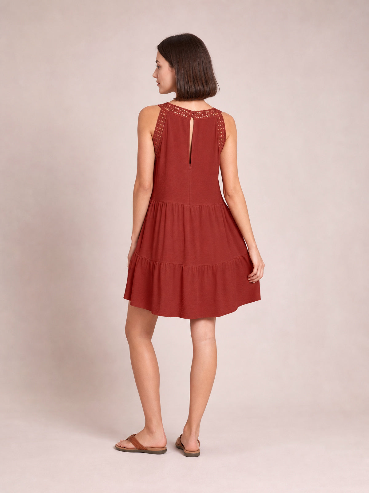 Model poses in a rust-red sleeveless lace dress with a relaxed tiered silhouette, gathered skirt, ruffled hem, and back keyhole detail.