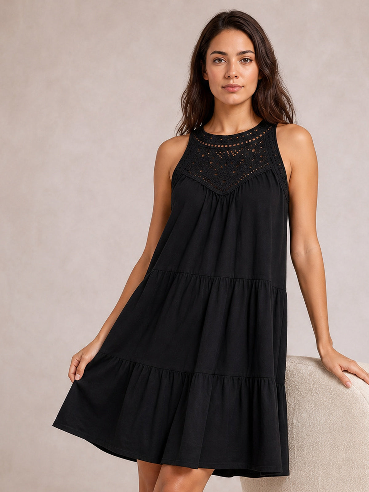 A model holds the hem of a black lace sleeveless dress with a relaxed, tiered silhouette, gathered construction, and gently flared ruffled skirt.