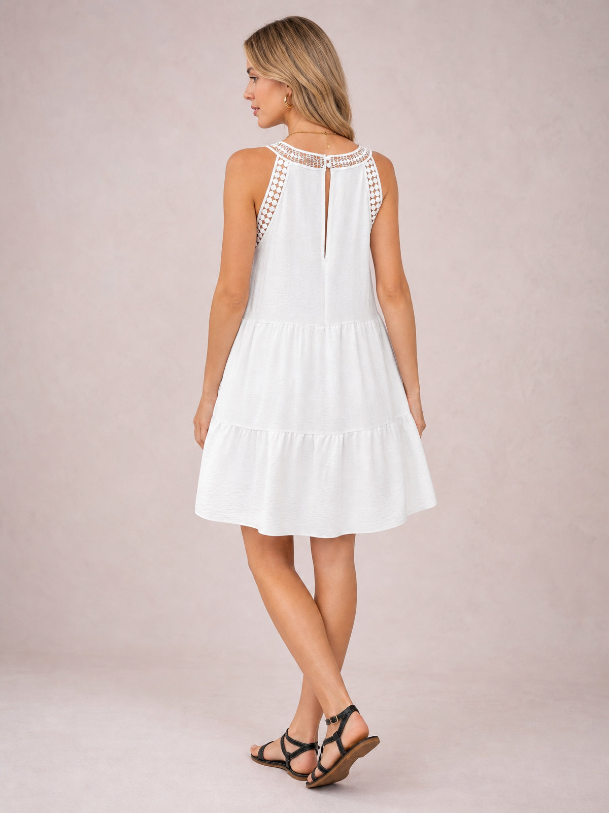 A model poses with crossed legs in a white sleeveless lace dress featuring a lace-trimmed neckline and shoulders, back keyhole opening, and tiered ruffled hem.