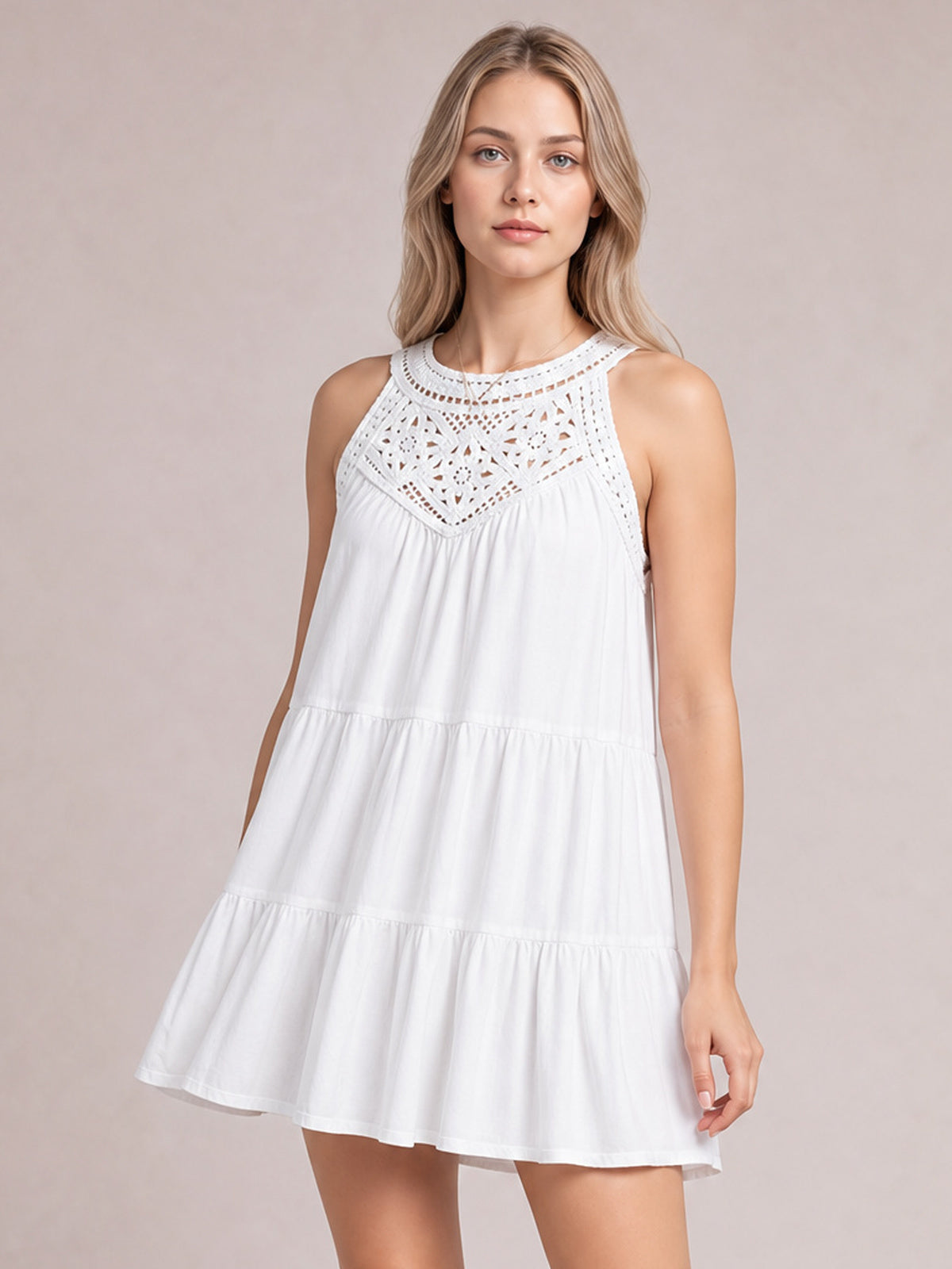 A model stands in a white sleeveless lace dress with a high neckline, intricate bodice, relaxed tiered silhouette, and gently flared ruffled hem.