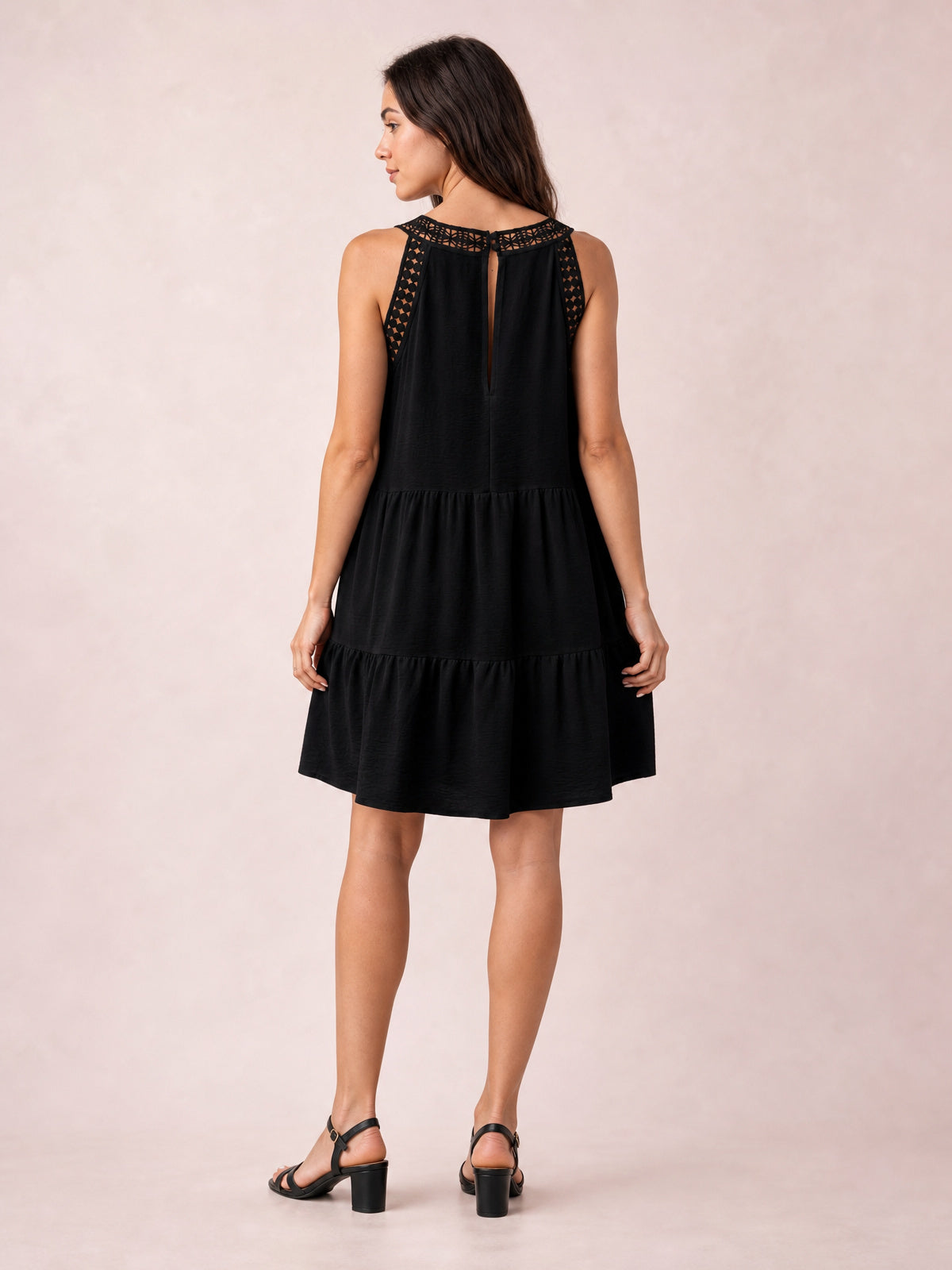 A model shows the back of a black sleeveless lace dress with a buttoned keyhole neckline, relaxed tiered silhouette, gathered seams, and a ruffled hem.