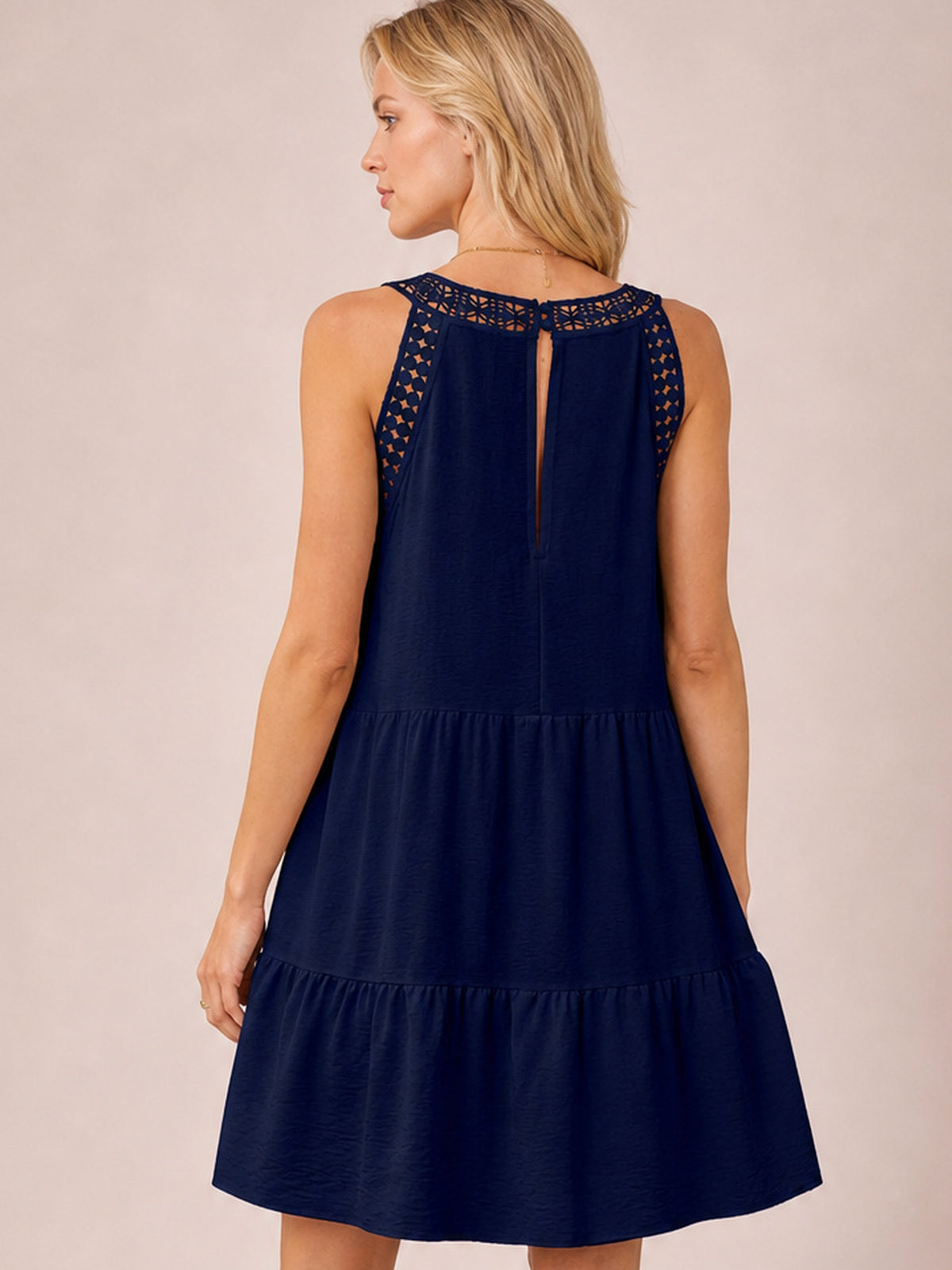 Model shown from the back in a navy sleeveless lace dress with a buttoned keyhole closure, relaxed tiered silhouette, and flared ruffled hem.