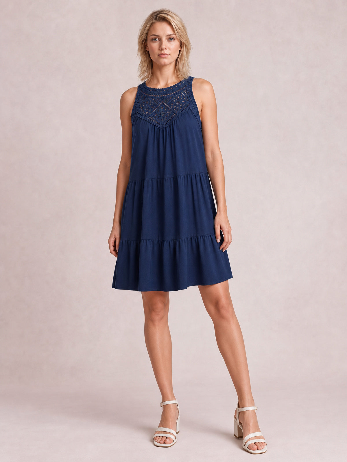 Model wears a navy sleeveless lace dress with a relaxed tiered silhouette, gathered skirt, intricate bodice detailing, and ruffled hem while standing with one leg angled.