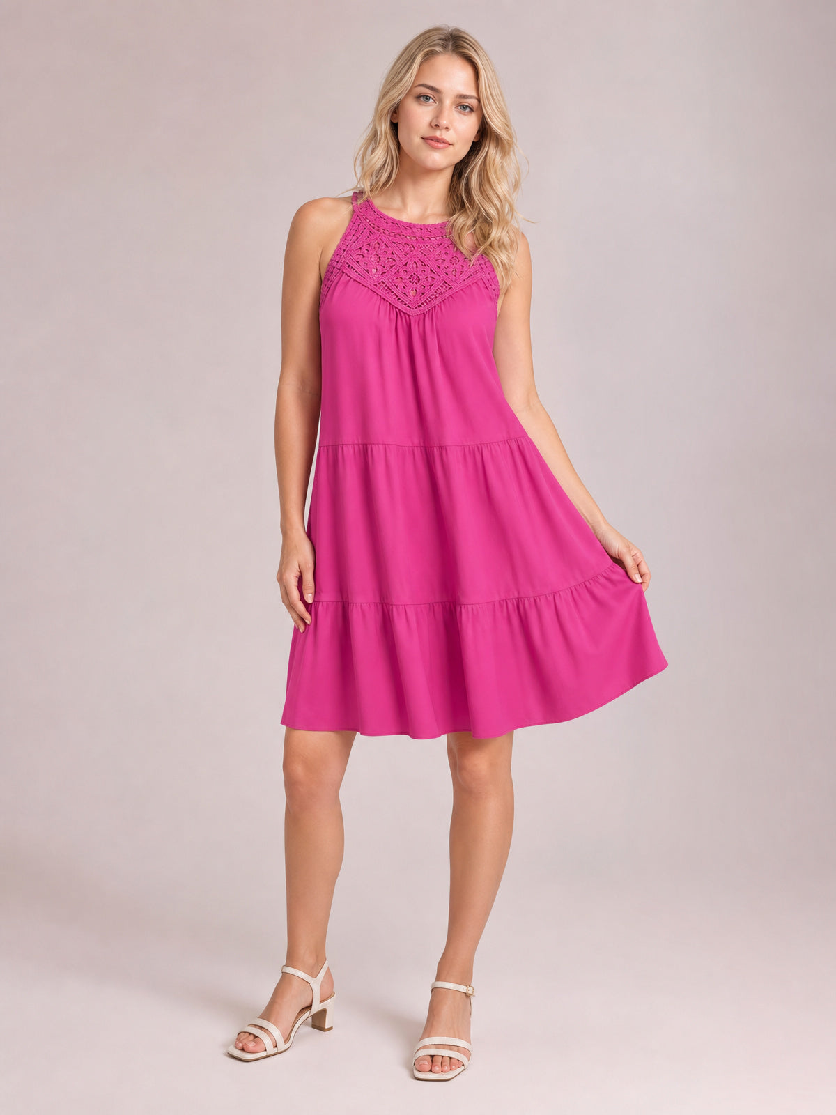 A model stands in a bright pink sleeveless lace dress with a relaxed, tiered silhouette, gathered skirt, and ruffled hem, holding the skirt out to show its flare.