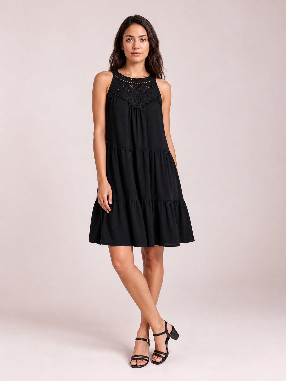 Model in a relaxed pose wears a black sleeveless lace dress with a tiered silhouette, gathered seams, buttoned lace bodice, and ruffled hem.