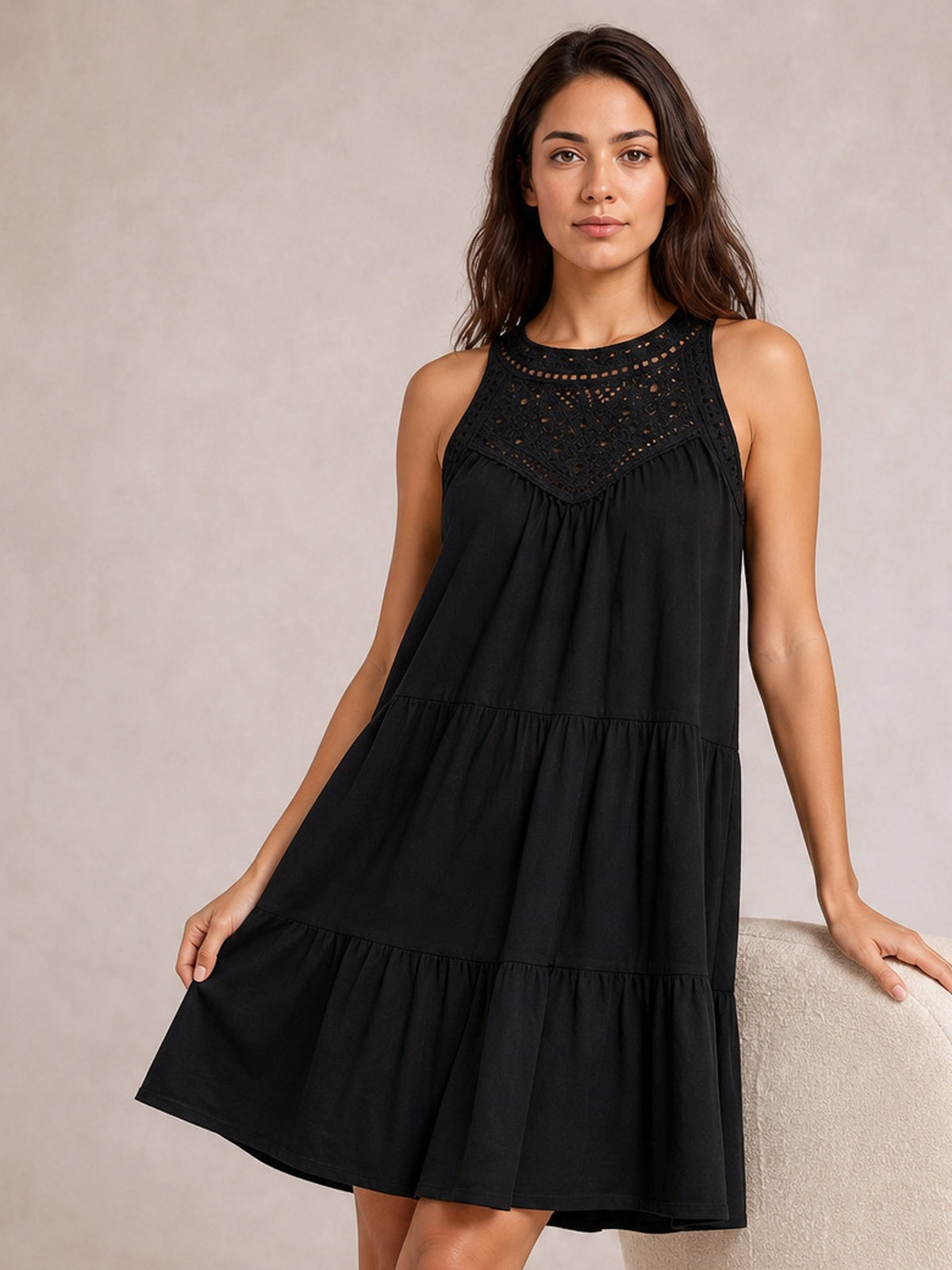 A model holds the hem of a black lace sleeveless dress with a relaxed, tiered silhouette, gathered construction, and gently flared ruffled skirt.