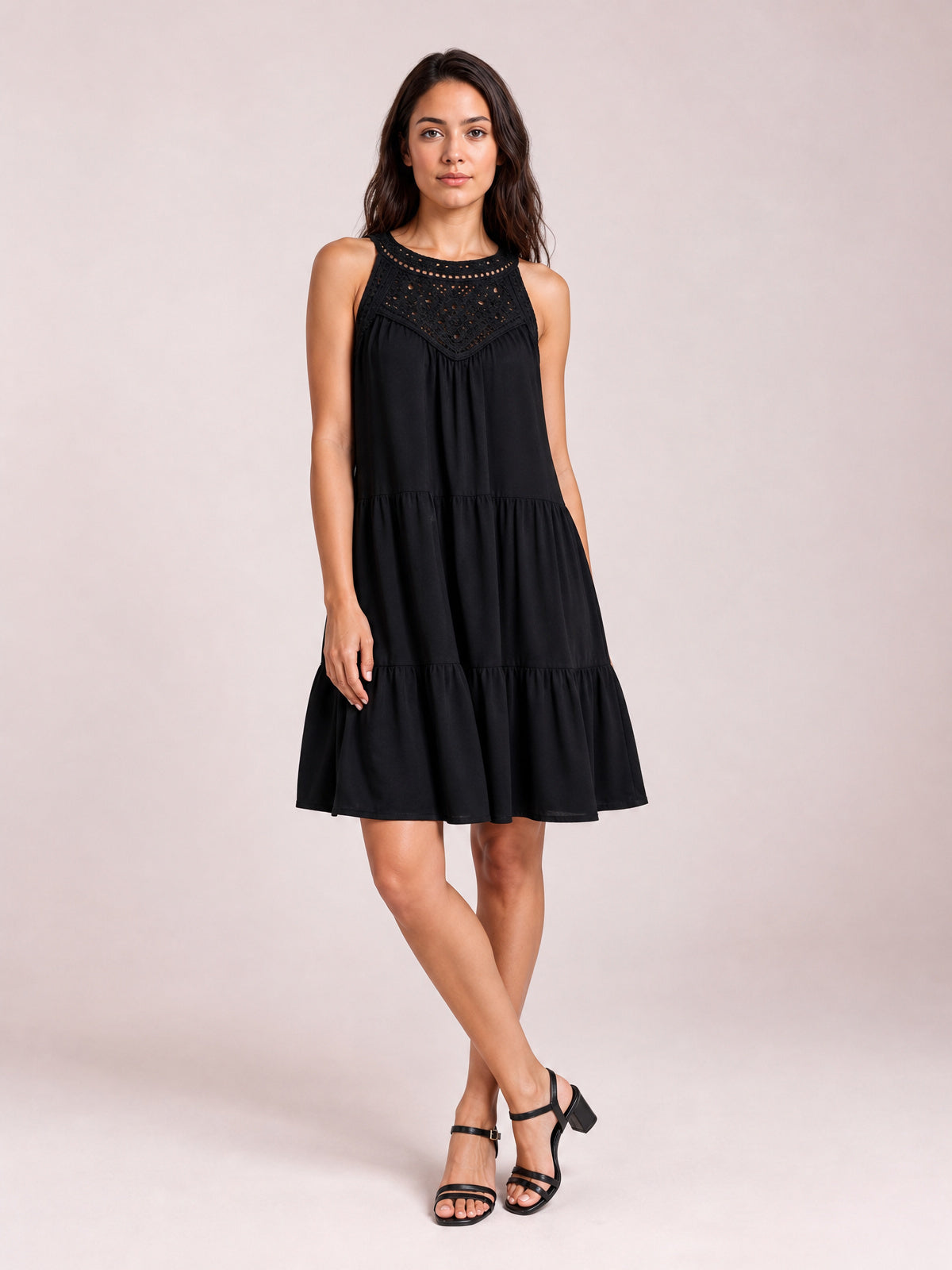 Model in a relaxed pose wears a black sleeveless lace dress with a tiered silhouette, gathered seams, buttoned lace bodice, and ruffled hem.
