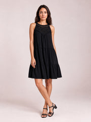 Model in a relaxed pose wears a black sleeveless lace dress with a tiered silhouette, gathered seams, buttoned lace bodice, and ruffled hem.