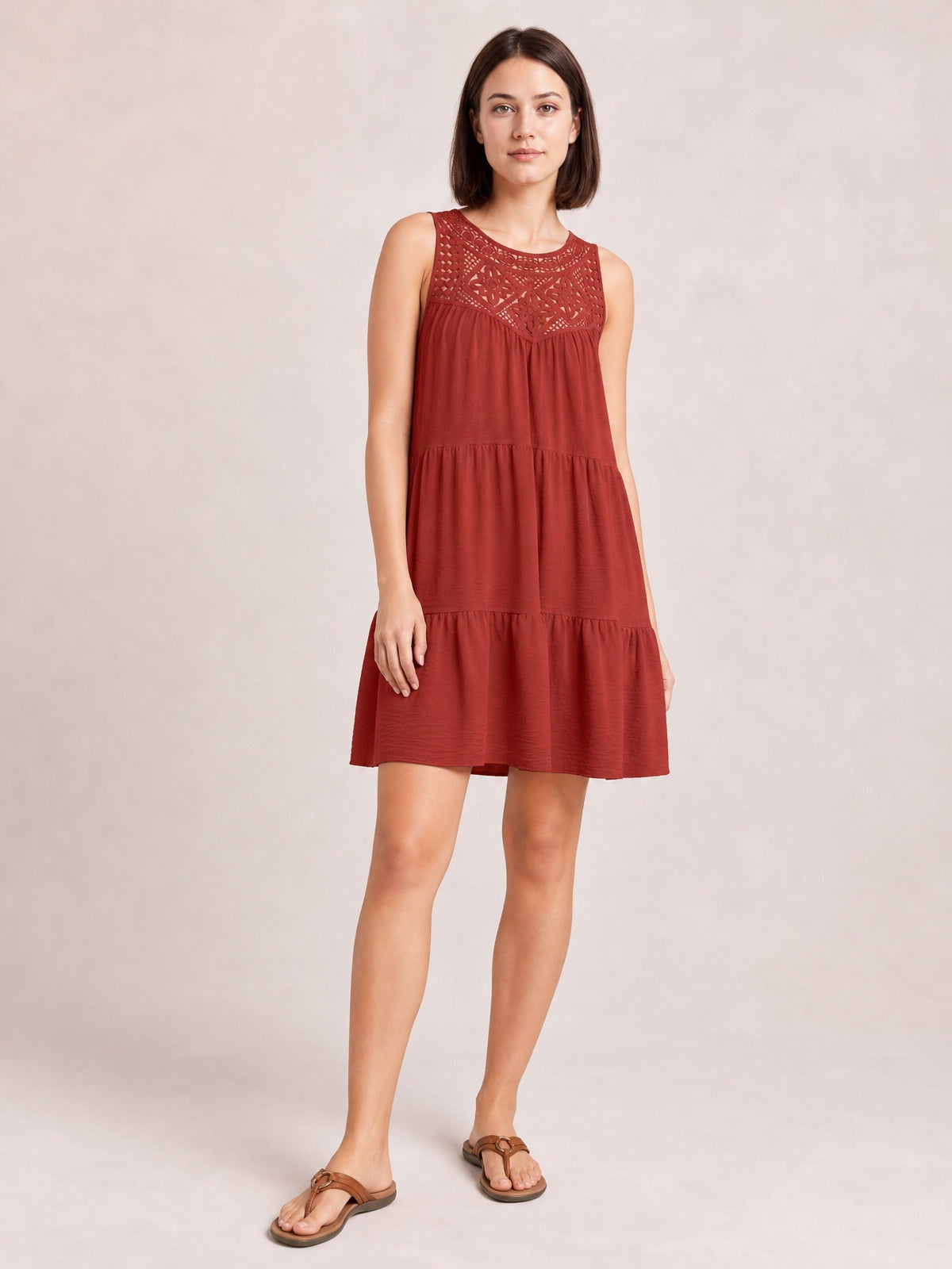 Model stands in a rust-red sleeveless lace dress with an intricate bodice, relaxed tiered silhouette, gathered skirt, and gently ruffled hem.