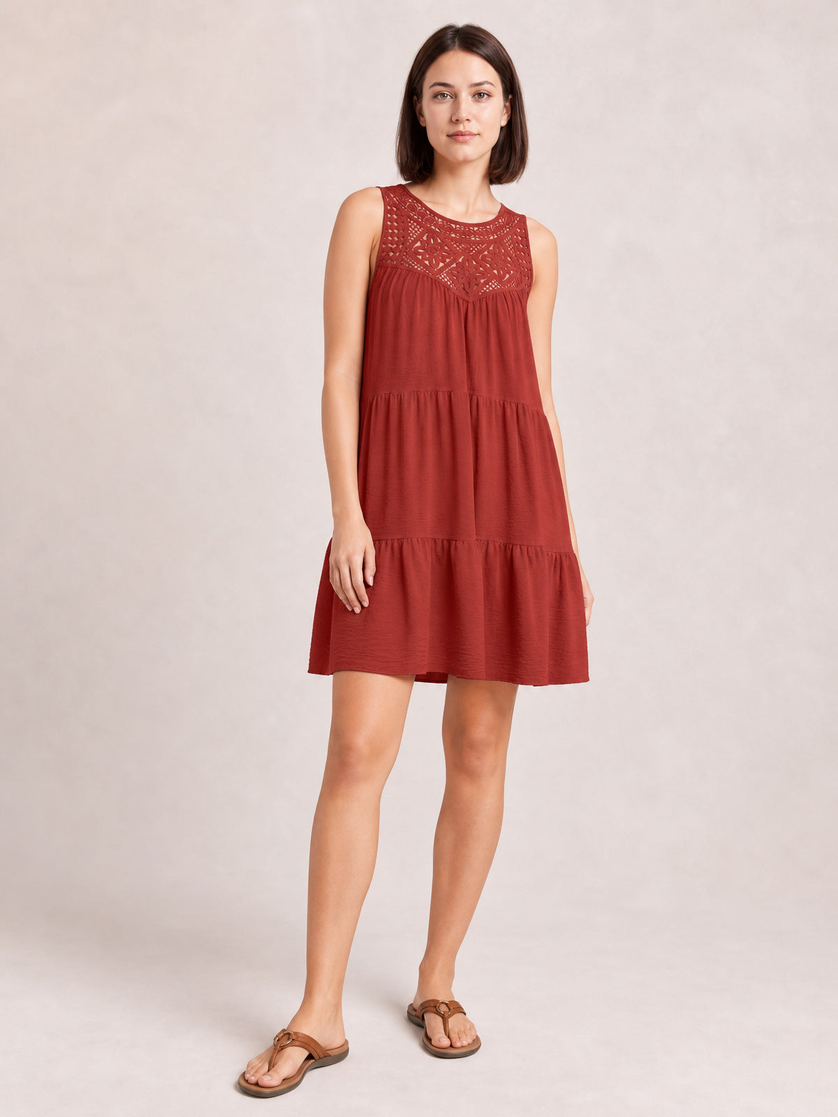 Model stands in a rust-red sleeveless lace dress with an intricate bodice, relaxed tiered silhouette, gathered skirt, and gently ruffled hem.