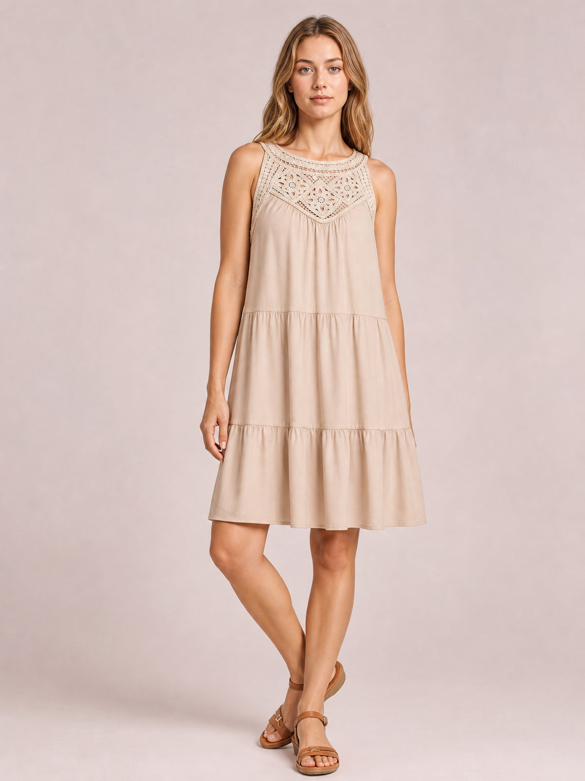 Model poses in a beige sleeveless lace dress with a relaxed tiered silhouette, gathered skirt panels, and a softly flared ruffled hem.