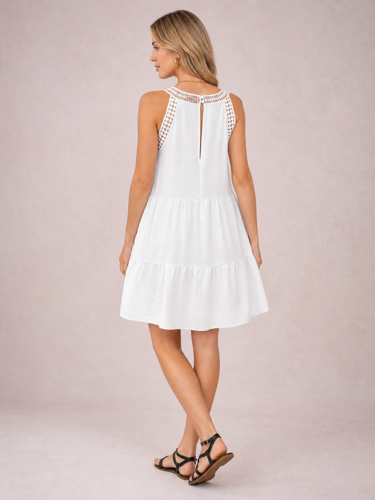 A model poses with crossed legs in a white sleeveless lace dress featuring a lace-trimmed neckline and shoulders, back keyhole opening, and tiered ruffled hem.