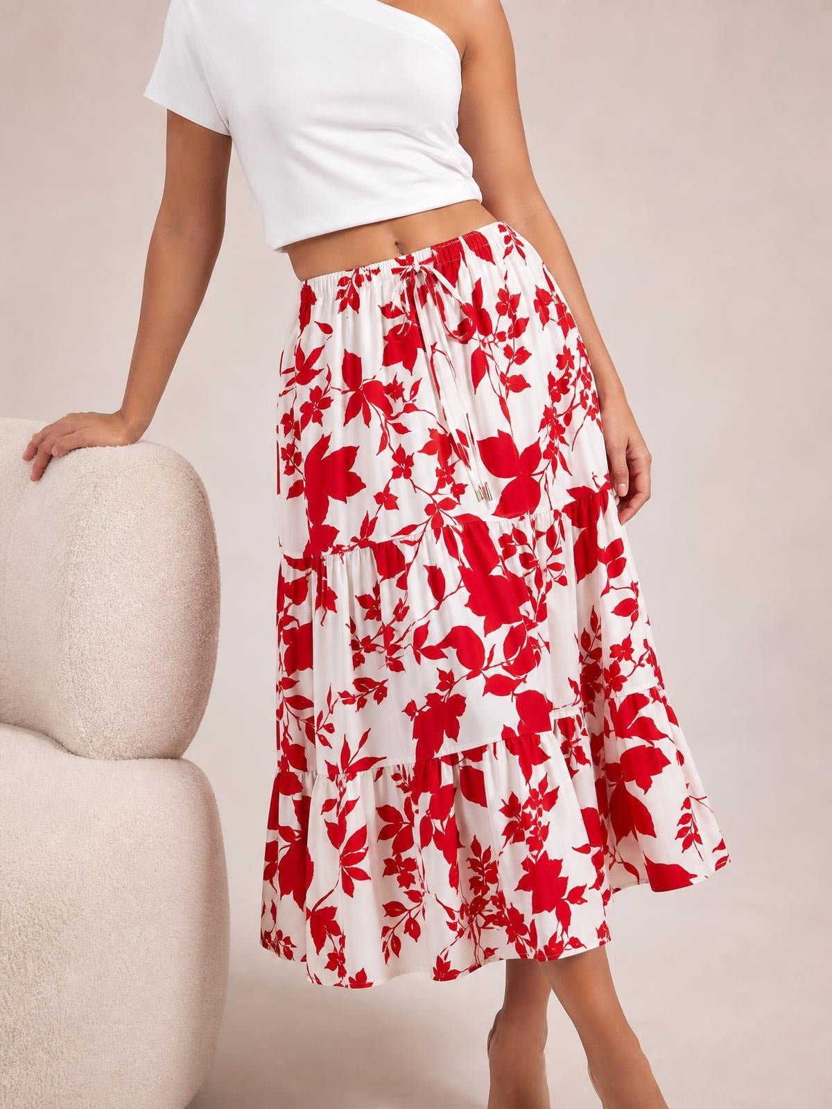 Model wears a flowy white midi skirt with bold red floral print, elastic drawstring waist, tiered construction, and ruffled hem while standing with one hand resting on a chair.