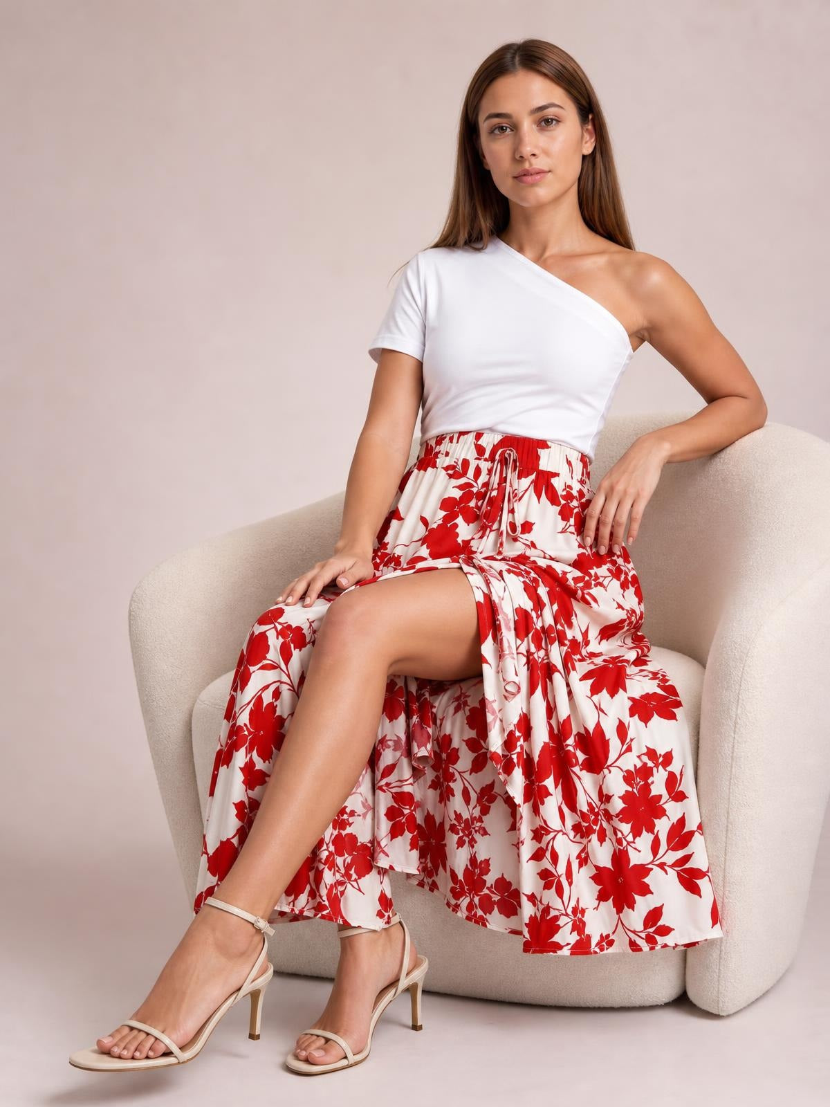 Seated model wears a flowy red-and-white floral printed midi skirt with a gathered waist, tie detail, and layered ruffled hem.