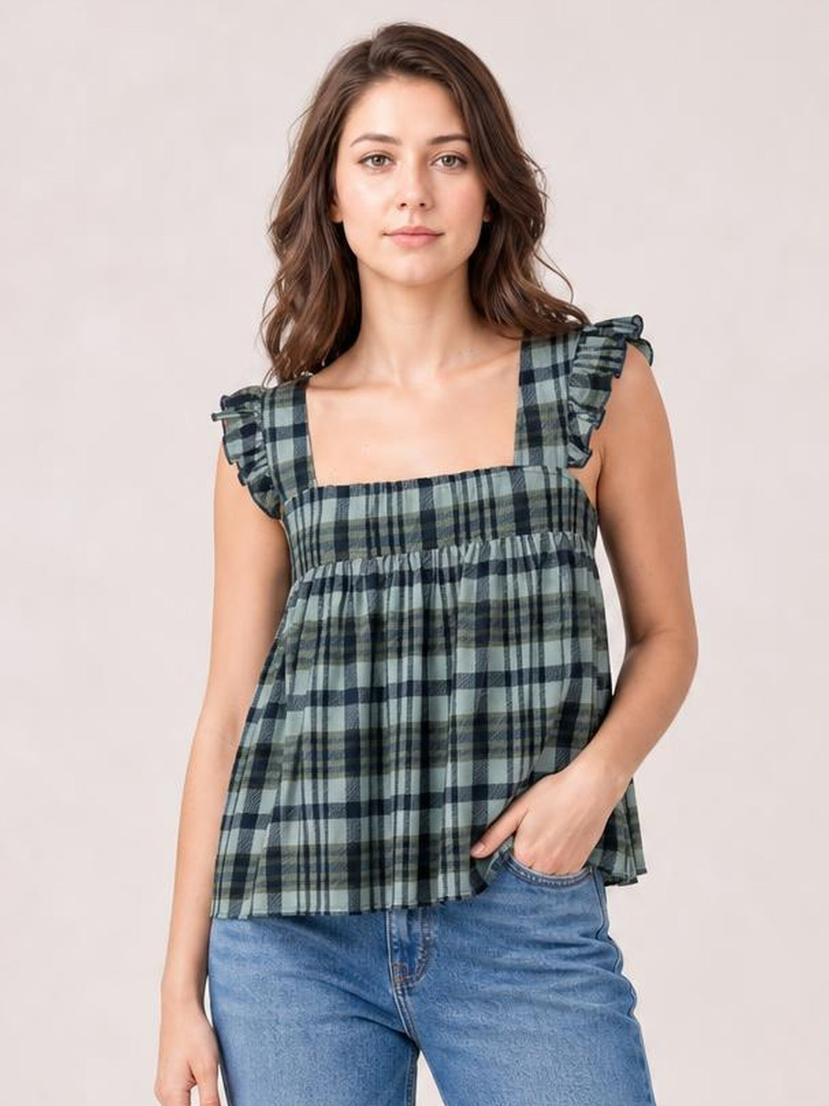Model wearing a blue-green plaid square neck tank with ruffled shoulder straps and a loose, tiered silhouette, standing with one hand in her jeans pocket.