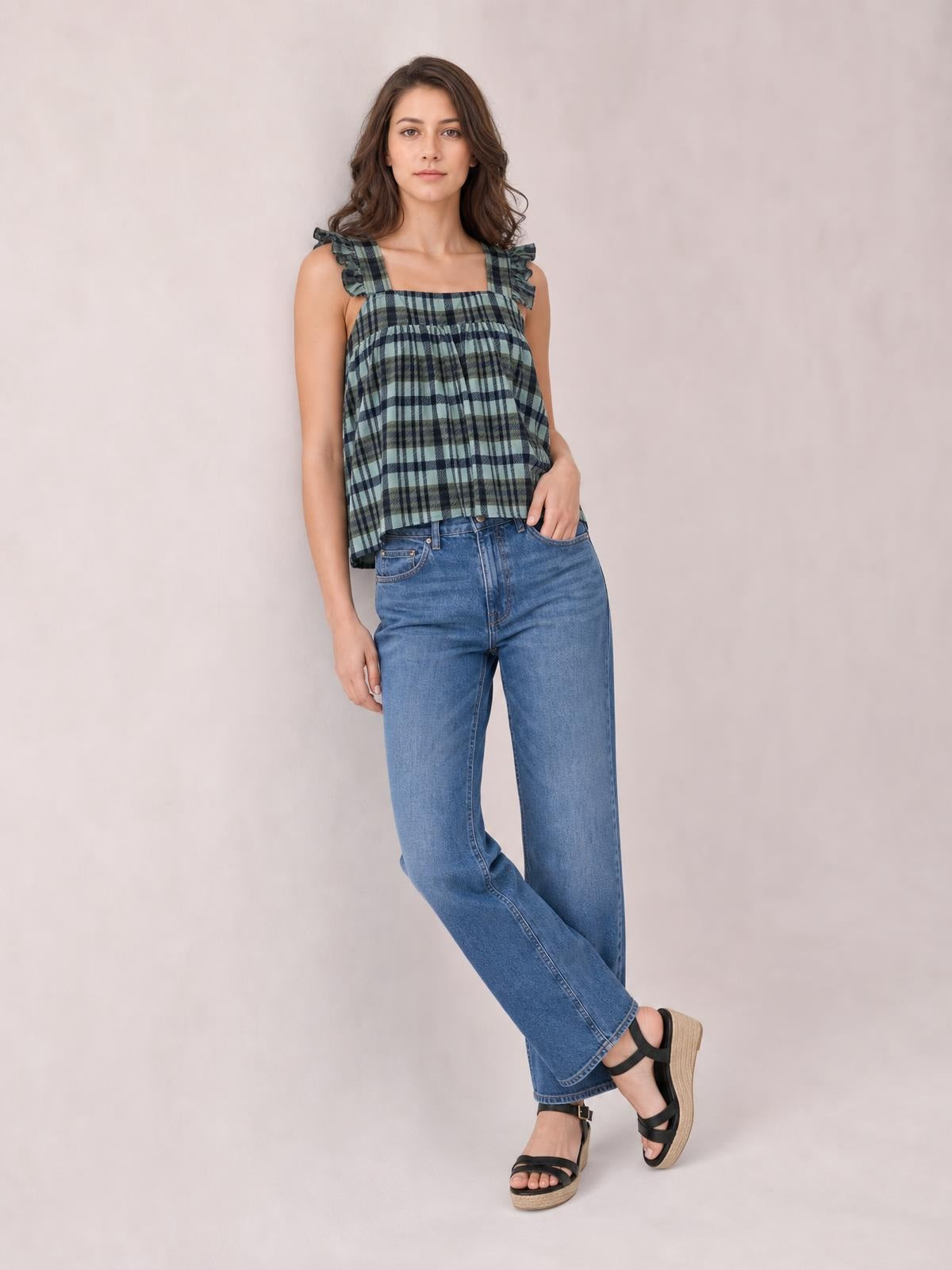 Model wears a green and navy plaid square neck tank with ruffled shoulder straps and a relaxed, gathered silhouette, posing in wide-leg jeans.