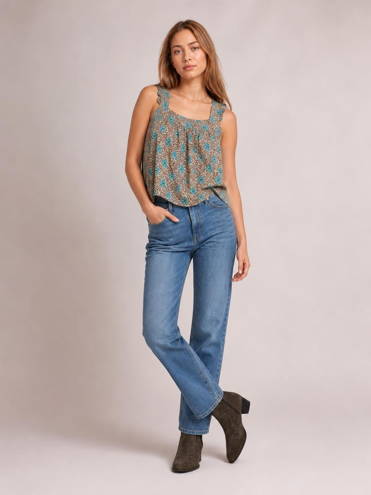 Model wears a taupe and teal printed tank with ruffled straps, a square neckline, and a relaxed, softly flowing silhouette, posing with one hand in her jeans pocket.