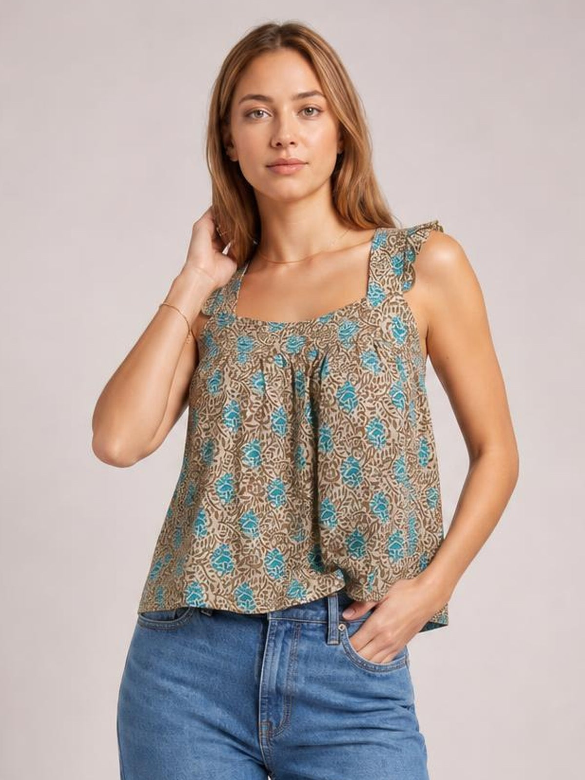 Model wears a brown and teal printed tank with a square neckline, ruffled shoulder straps, and a loose, flowy silhouette, posing with one hand in her pocket.