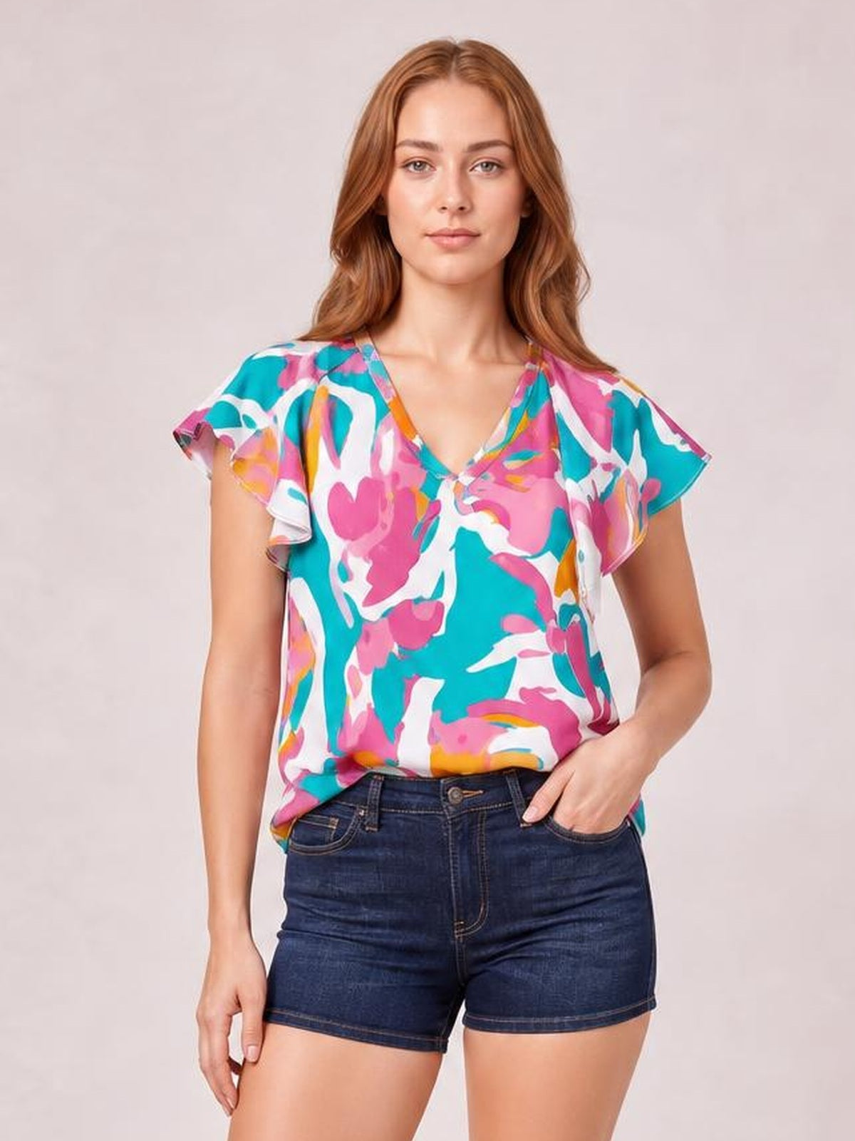 Model wearing a colorful printed blouse with ruffled cap sleeves, a notched V-neck, and a loose silhouette, posed with one hand in her denim shorts pocket.