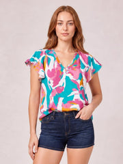 Model wearing a colorful printed blouse with ruffled cap sleeves, a notched V-neck, and a loose silhouette, posed with one hand in her denim shorts pocket.