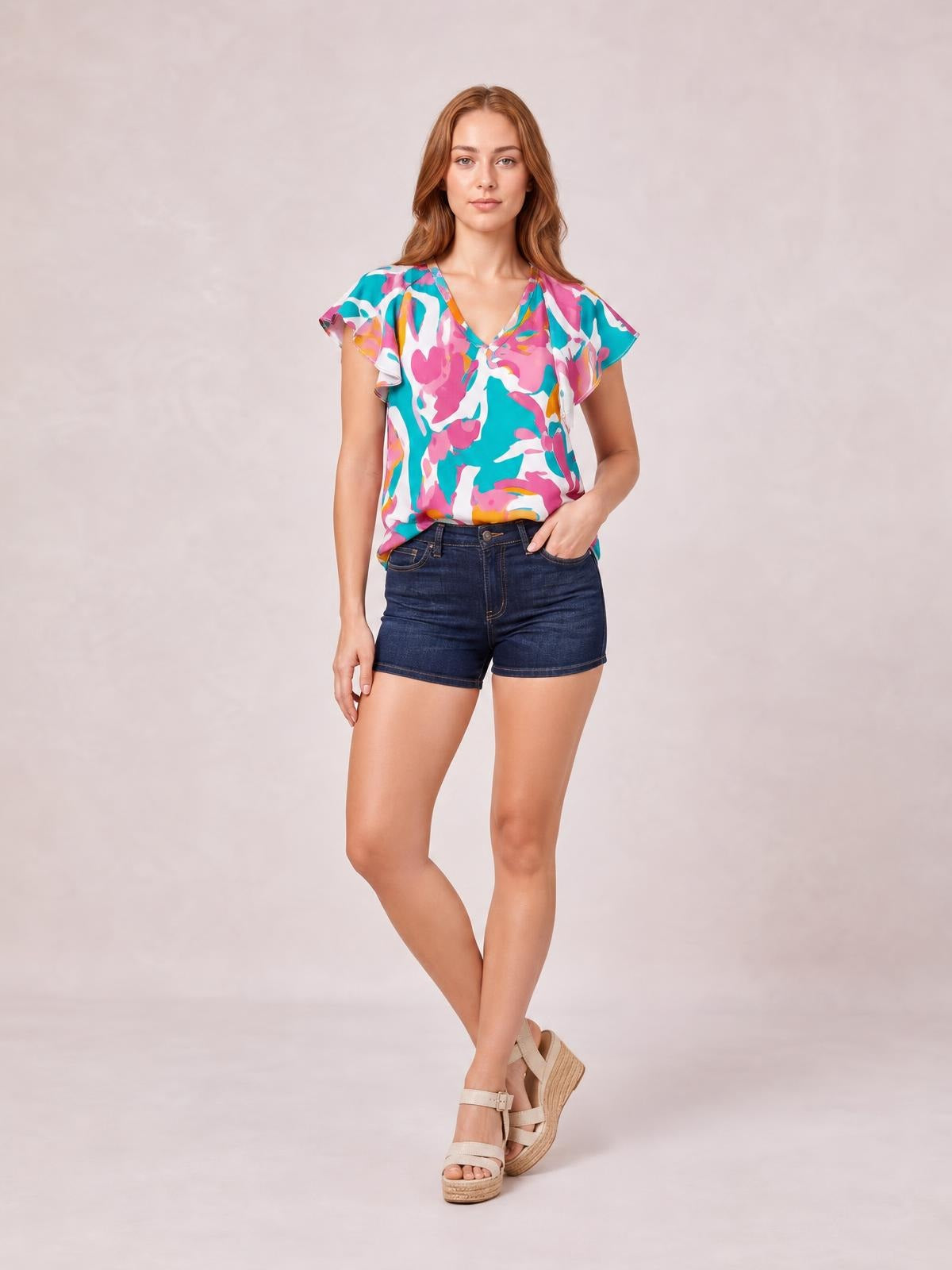 A model poses in a turquoise, pink, and white printed blouse with ruffled notched cap sleeves and a relaxed silhouette, one hand in her shorts pocket.