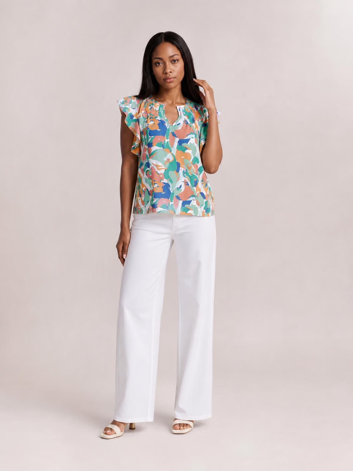 A model wears a colorful printed blouse with ruffled cap sleeves, a notched neckline, and a relaxed straight silhouette while touching her hair.