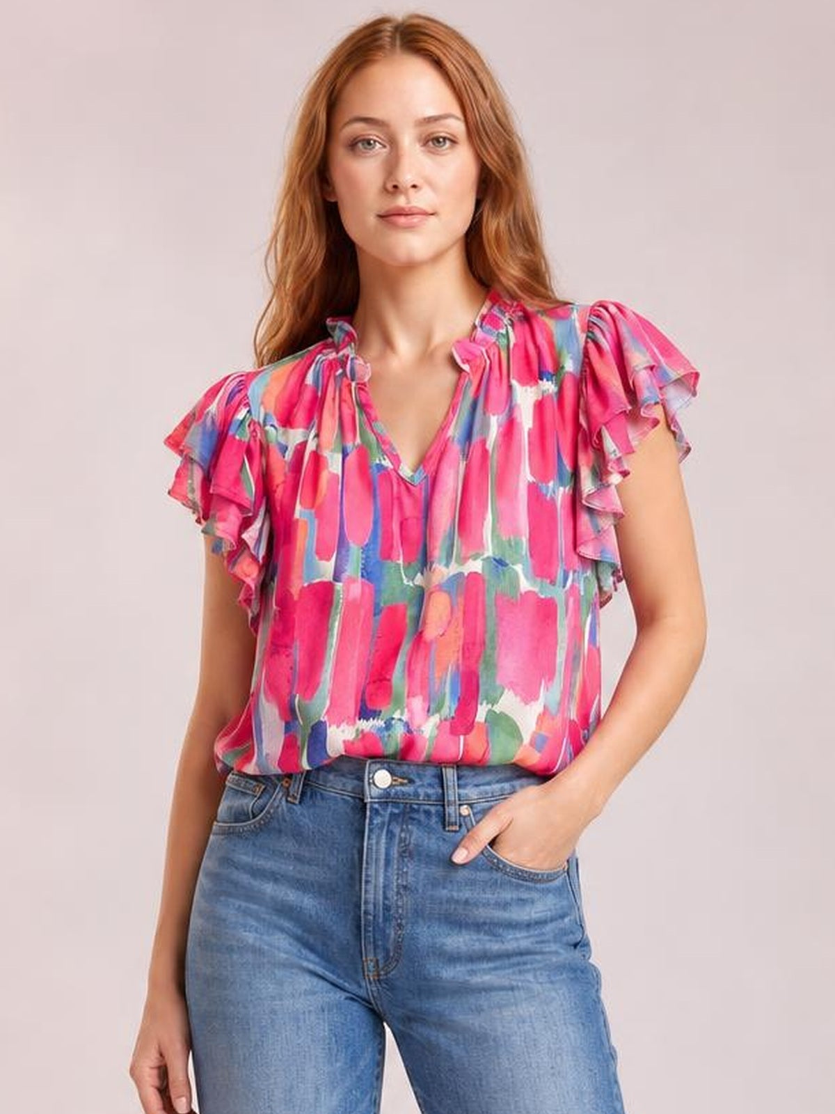 Model wearing a bright pink printed blouse with ruffled cap sleeves, a notched V-neck, and a relaxed silhouette, posed with one hand in her jeans pocket.